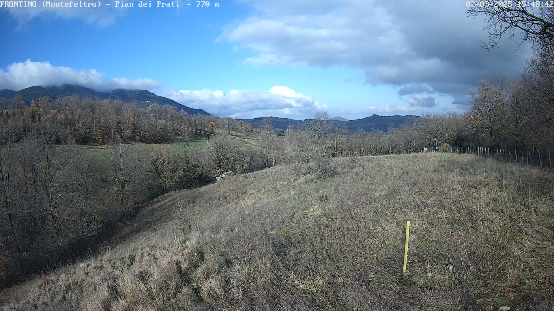 A partly cloudy day reveals a hilly landscape with leafless trees and dry, brown grass, mountains in the background.