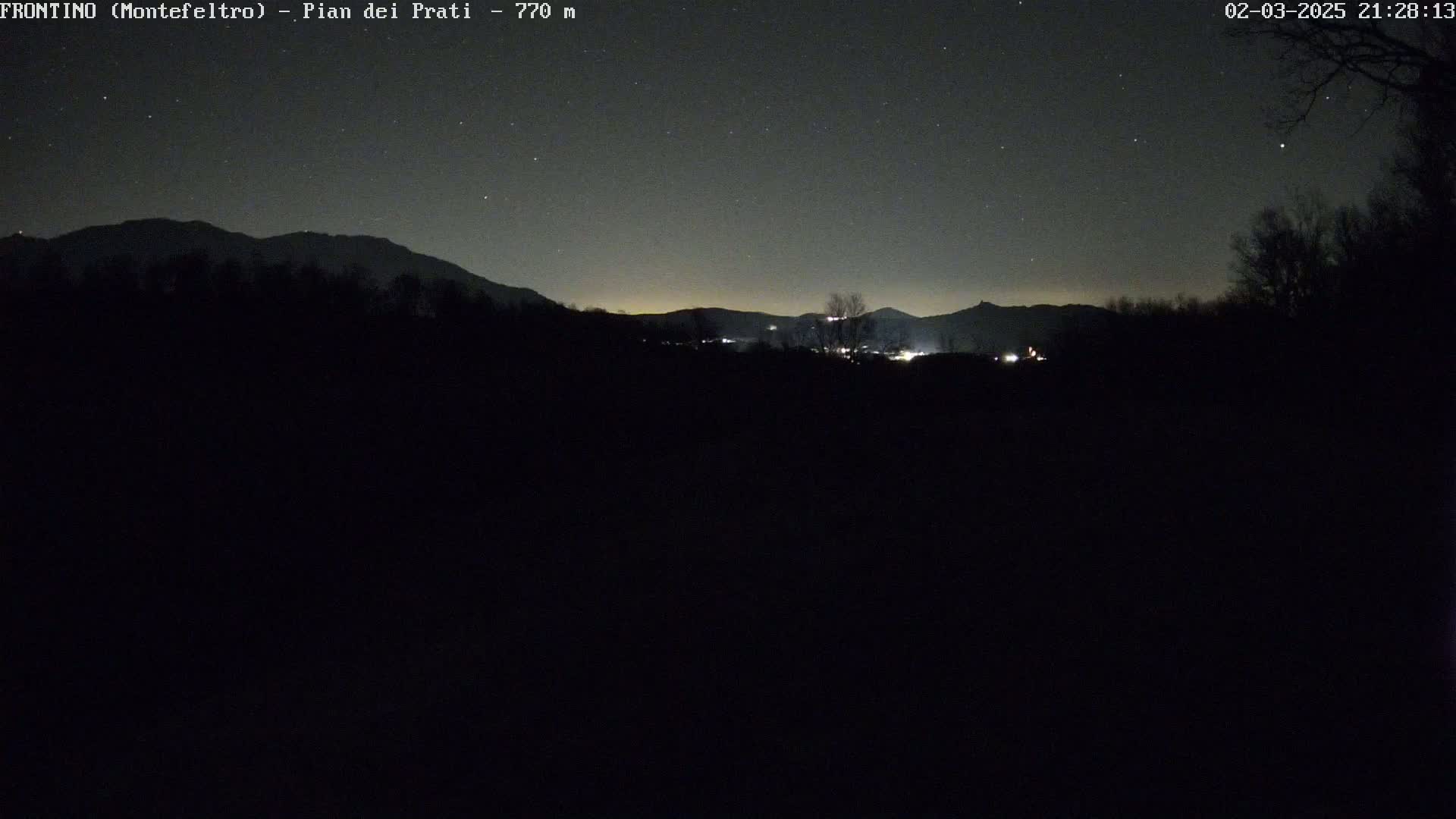 A nighttime, starlit view of a dark landscape featuring distant, dimly lit hills and mountains.