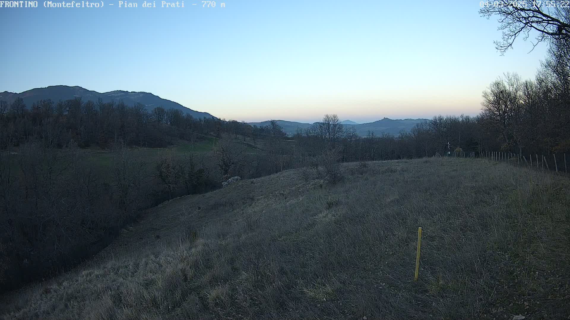 A hilly landscape at dusk shows a field of dry grass, bare winter trees, and distant mountains under a clear, pale blue sky.