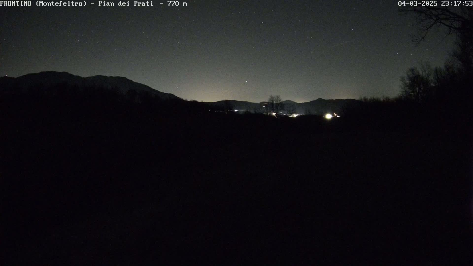 A nighttime view of dark mountains under a starry sky, with a few distant lights visible in the valley.