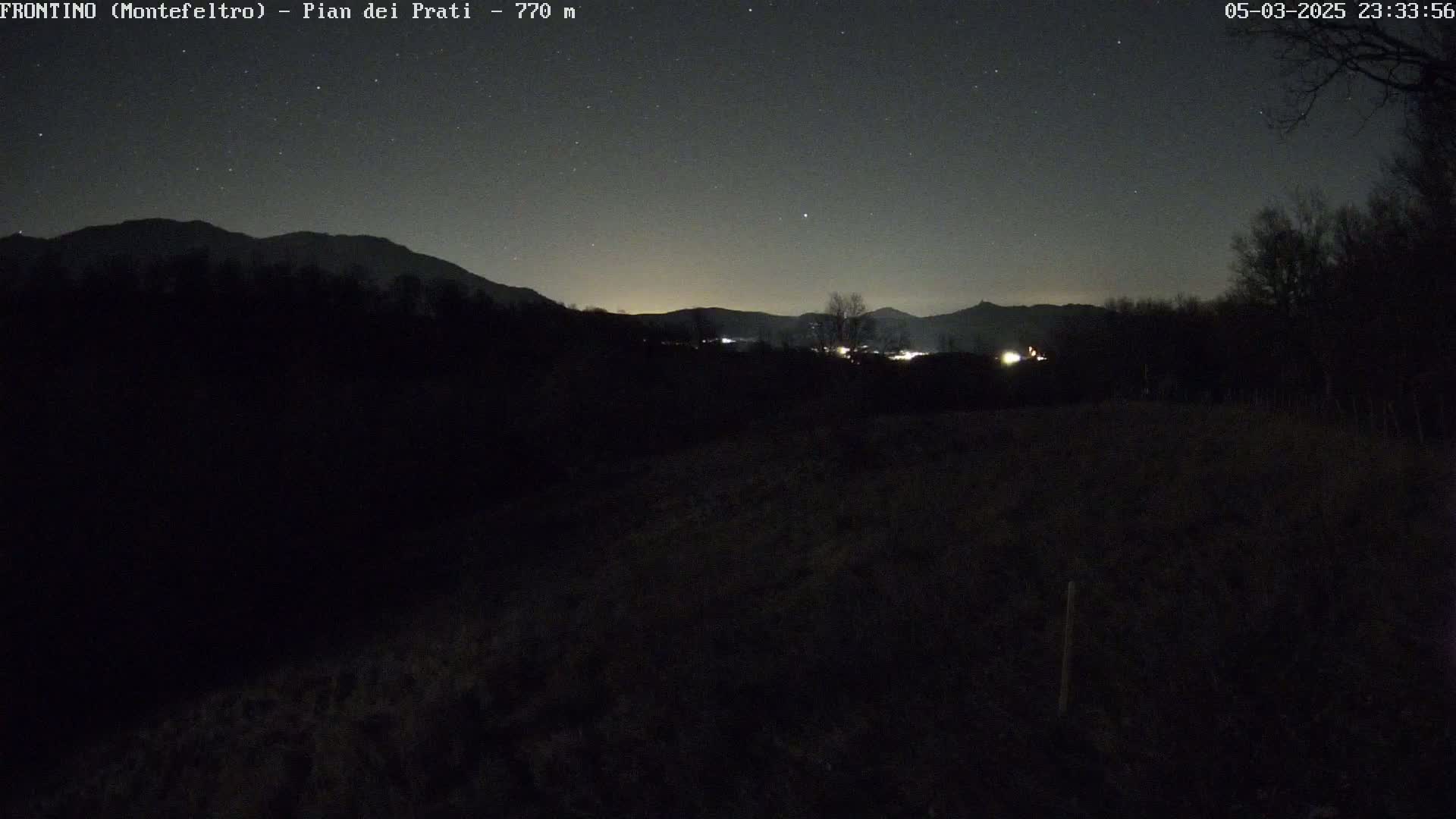 A nighttime landscape shows a dark field in the foreground, silhouetted trees and distant mountains under a starlit sky.