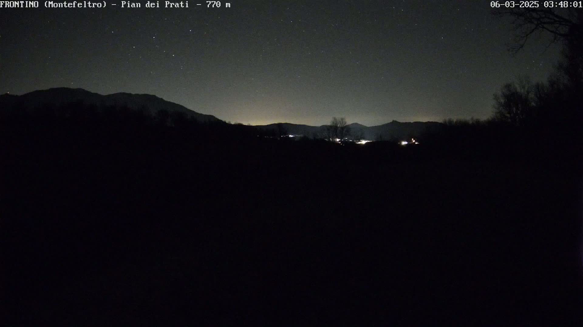 A nighttime, clear-sky view shows a dark, silhouetted landscape of mountains and trees, with a few faint lights visible in the distance.