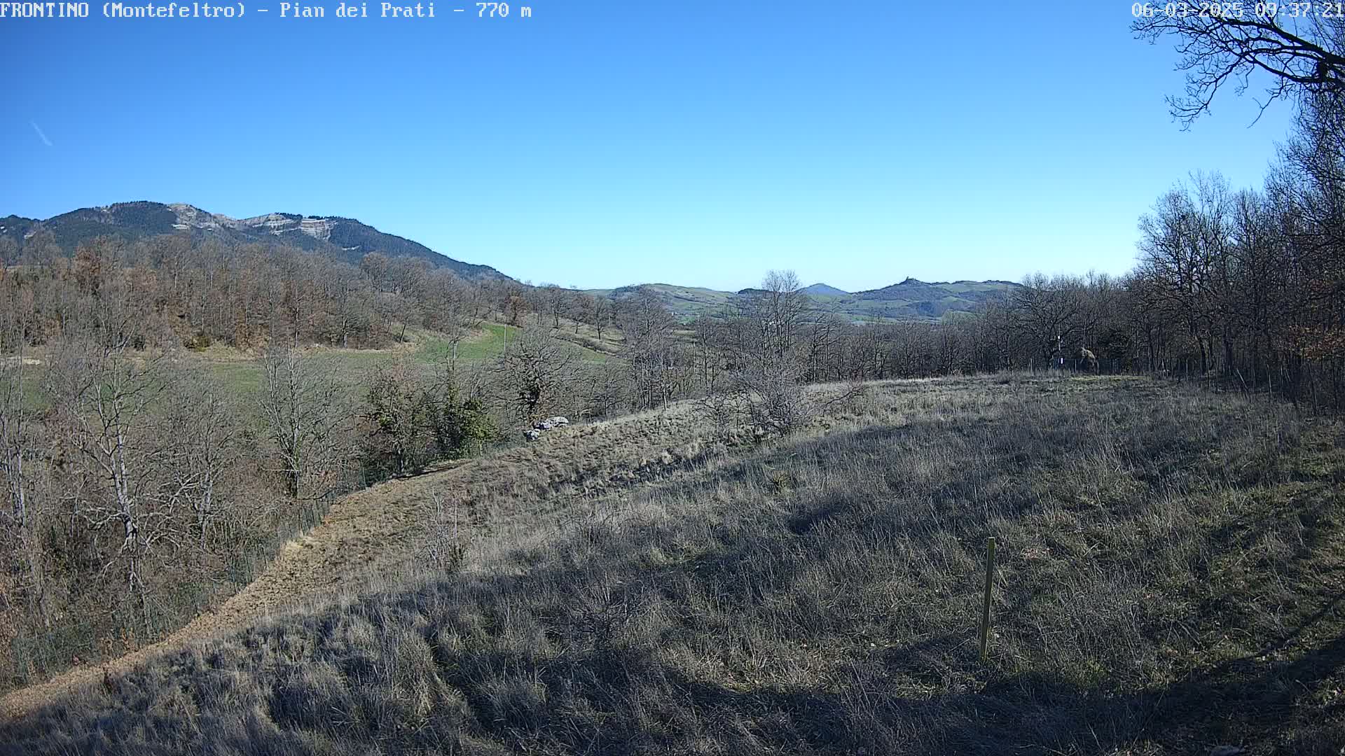 A sunlit, hilly landscape with bare, winter trees and dry, brown fields under a clear blue sky.