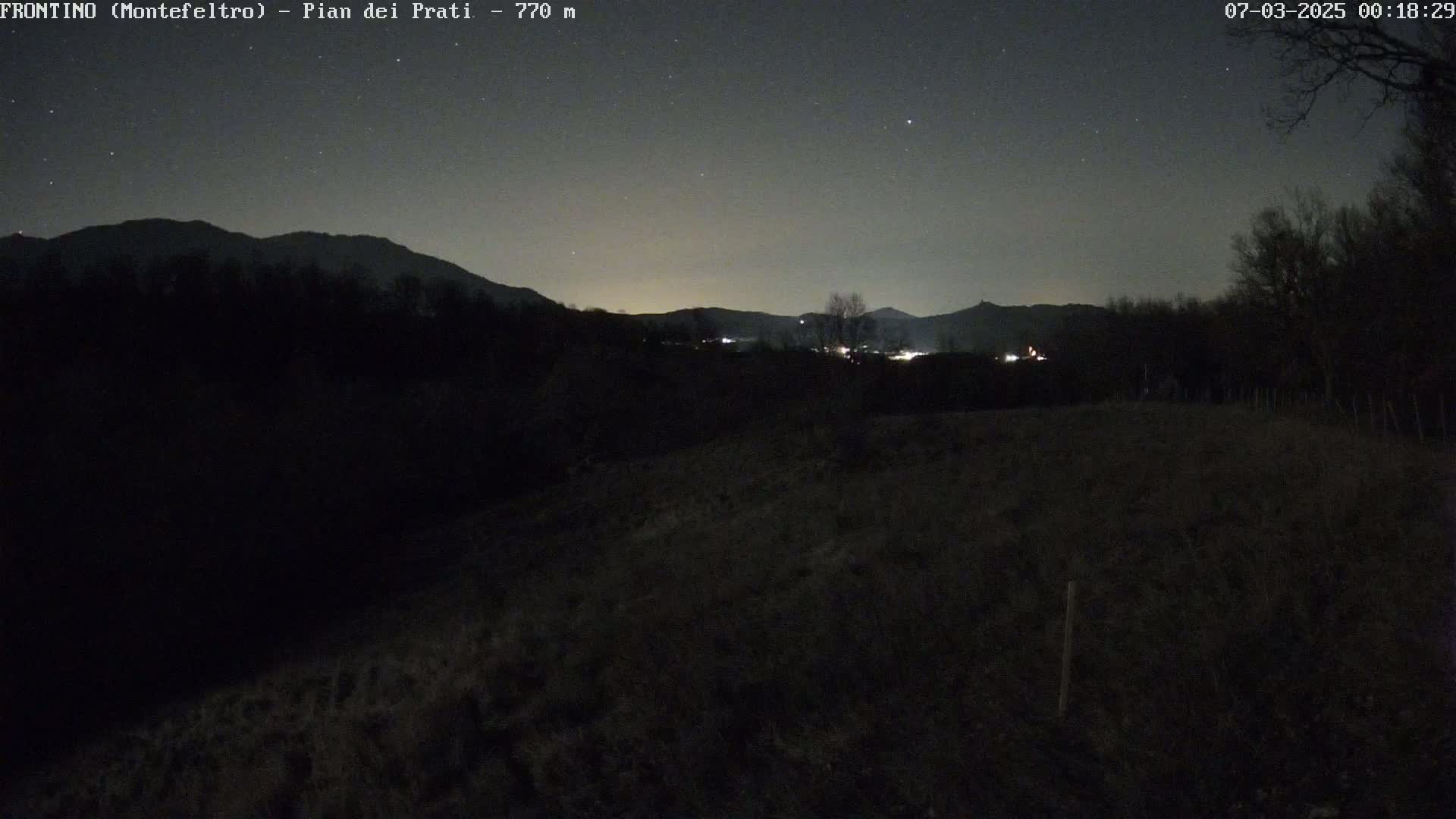 A nighttime, starlit view shows dark, silhouetted hills and trees, with a few distant lights visible in the valley.