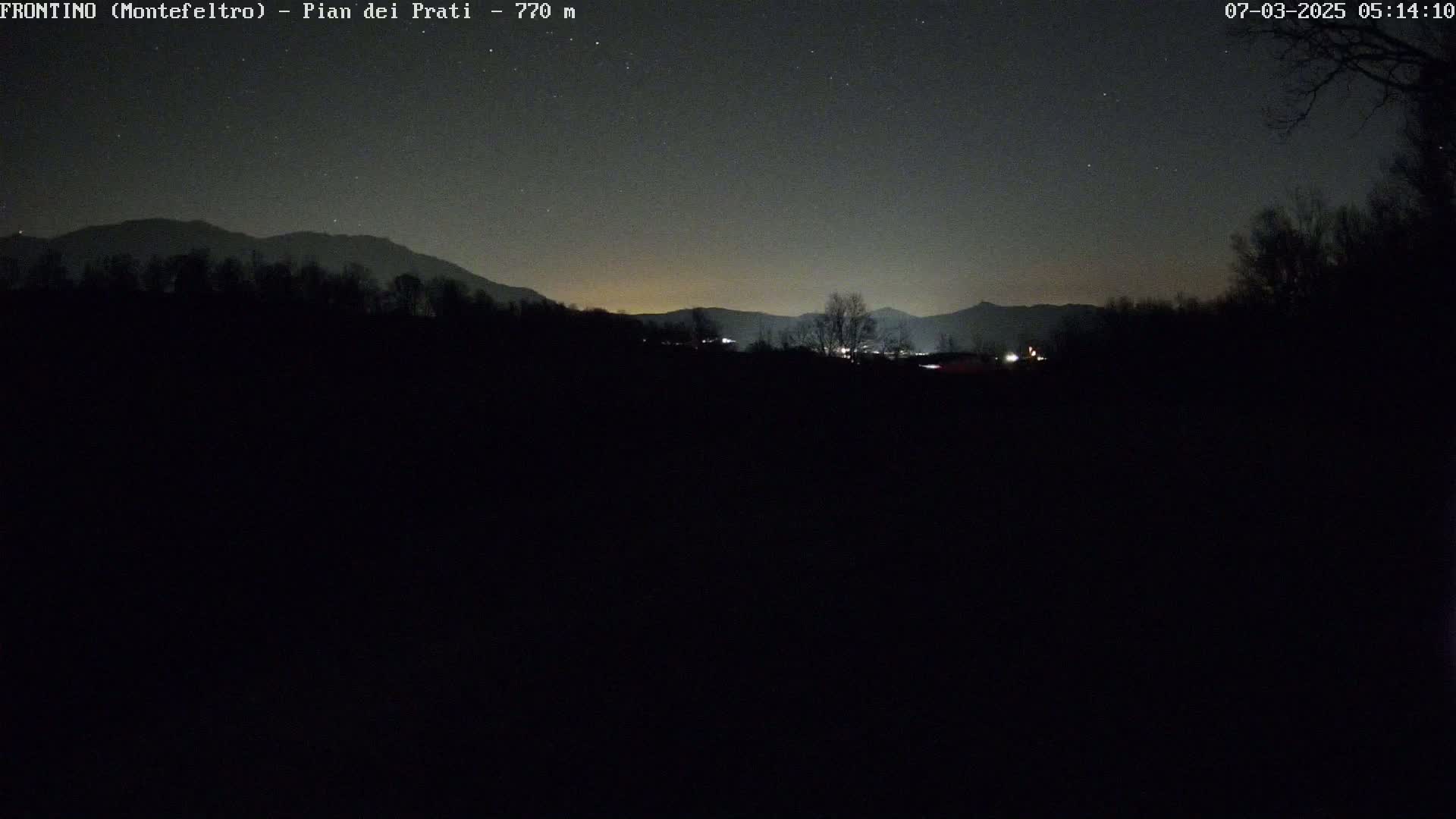 A nighttime view of a dark landscape featuring silhouetted trees and distant mountains under a clear, starry sky.