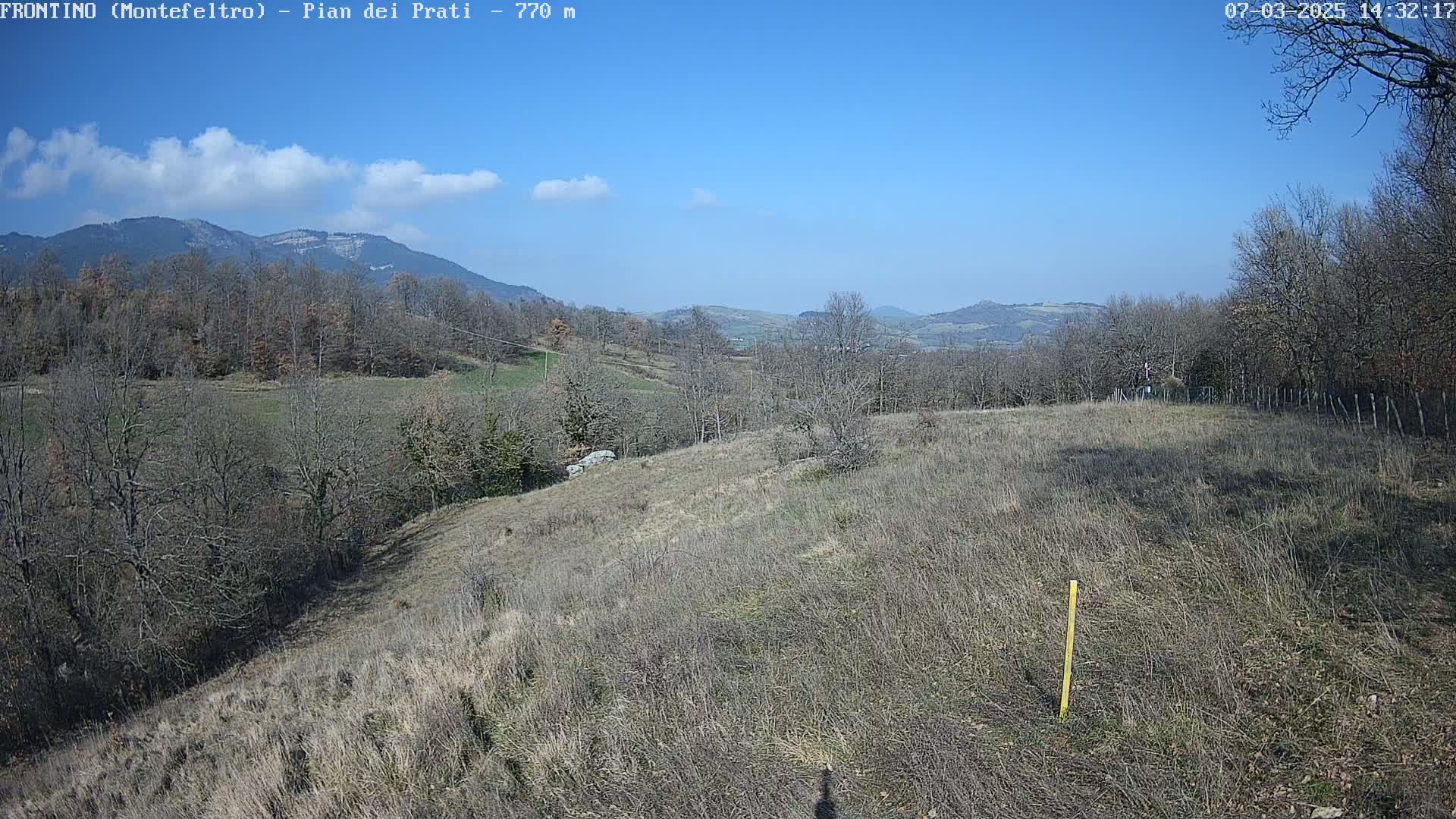 A sunny, clear day reveals a hilly landscape with bare, winter trees and dry, brown grass, extending to distant mountains under a bright blue sky.
