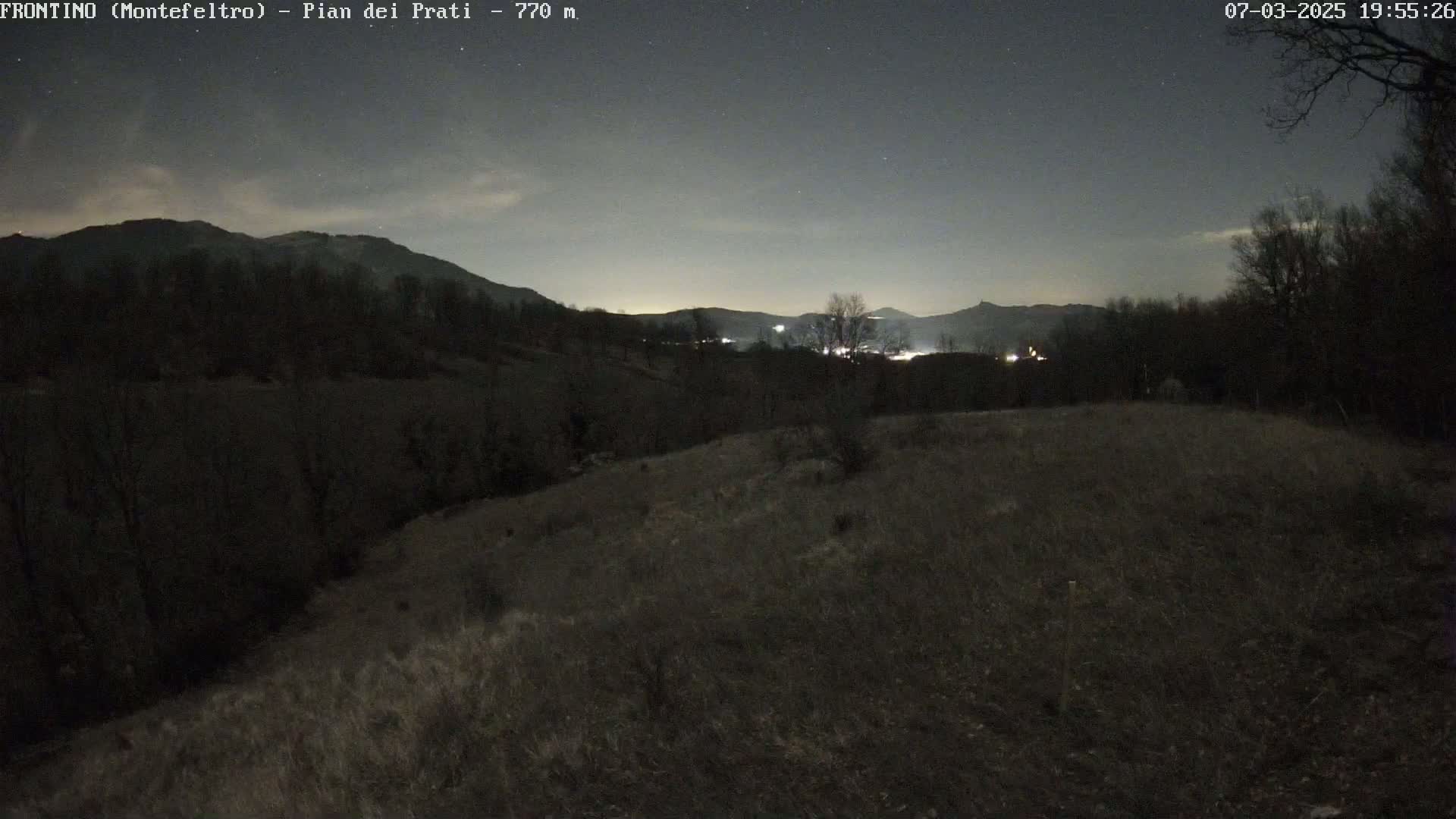 A nighttime view of a hilly, sparsely vegetated landscape under a clear, starry sky.