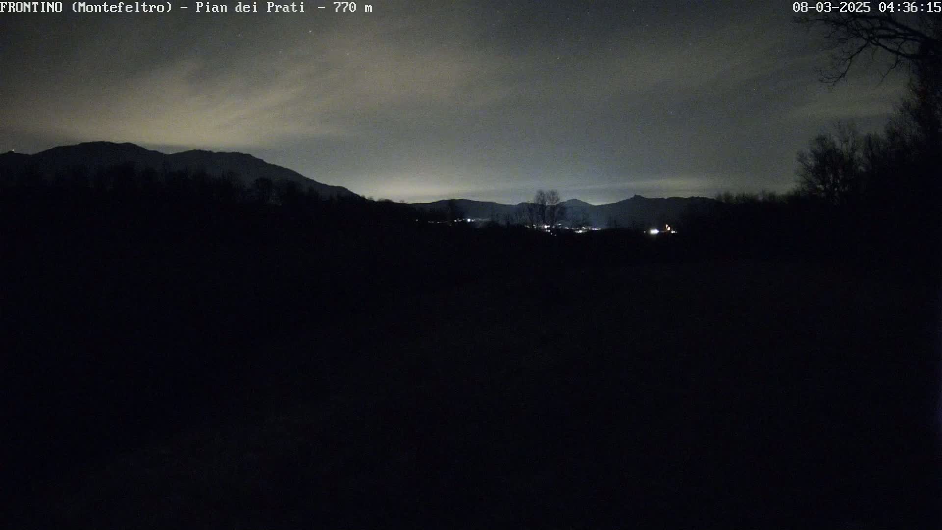 A nighttime view of dark, silhouetted mountains and trees under a mostly clear, dark sky.