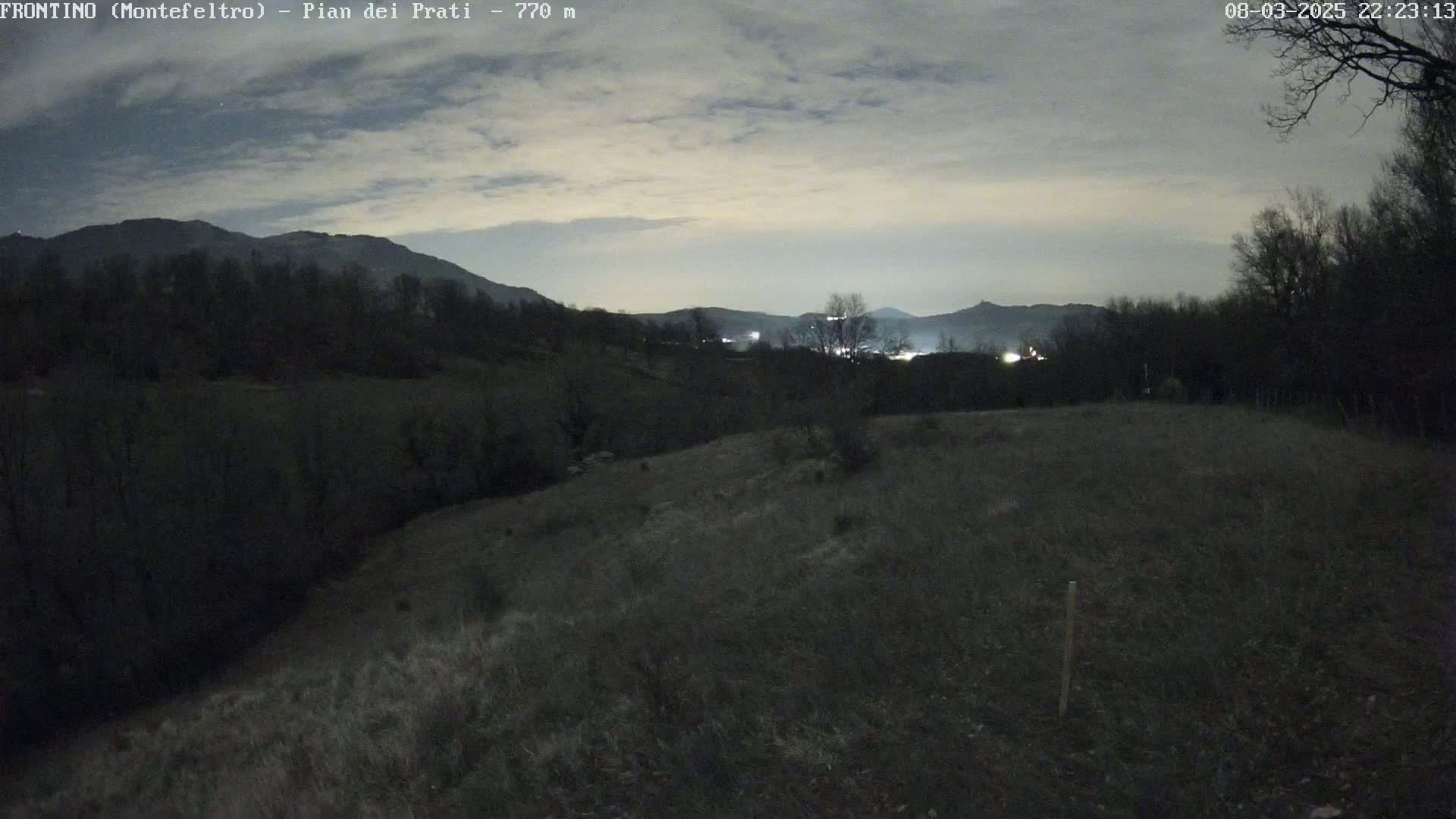A nighttime view of a grassy hillside overlooking a valley with distant lights and mountains under a cloudy sky.