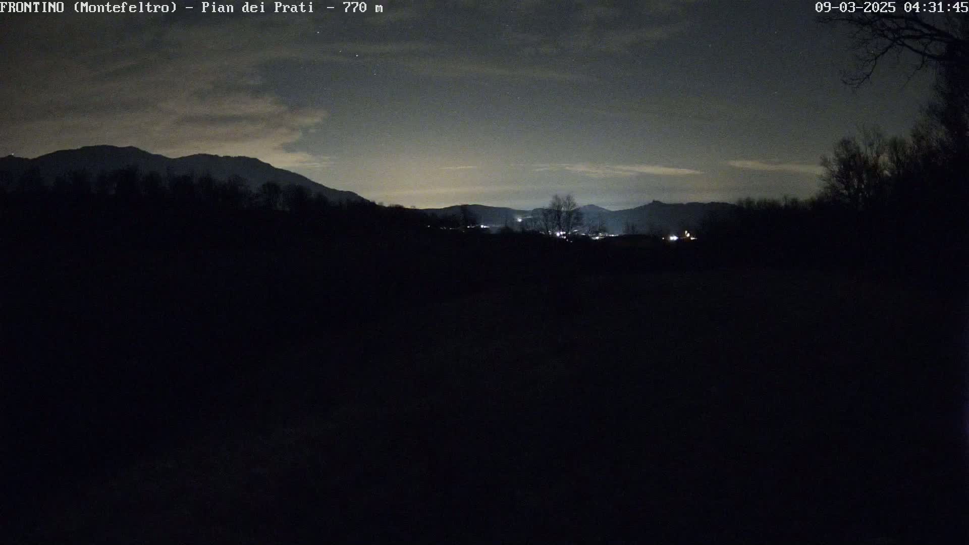 A nighttime view of silhouetted mountains and trees under a partly cloudy sky.