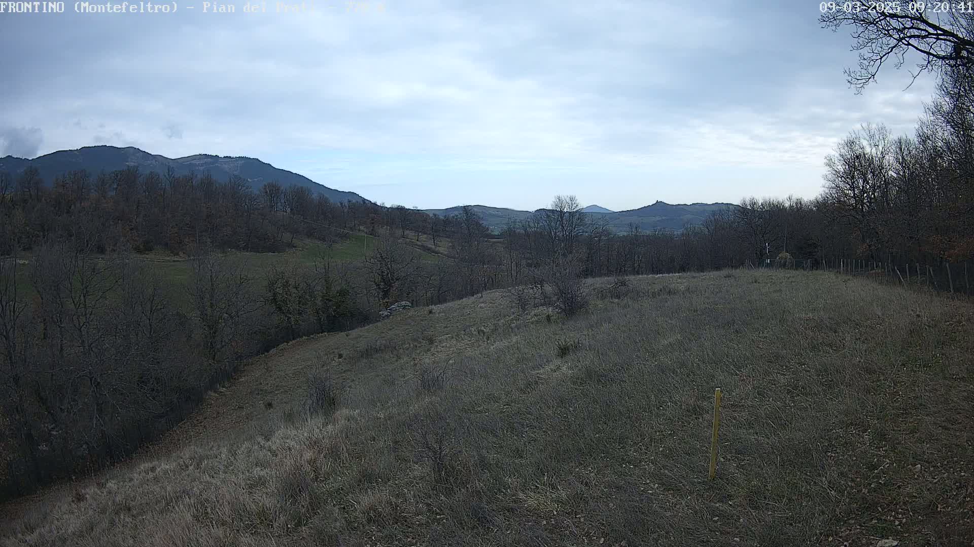 A view of a hilly, sparsely vegetated landscape under a cloudy sky, with distant mountains in the background.