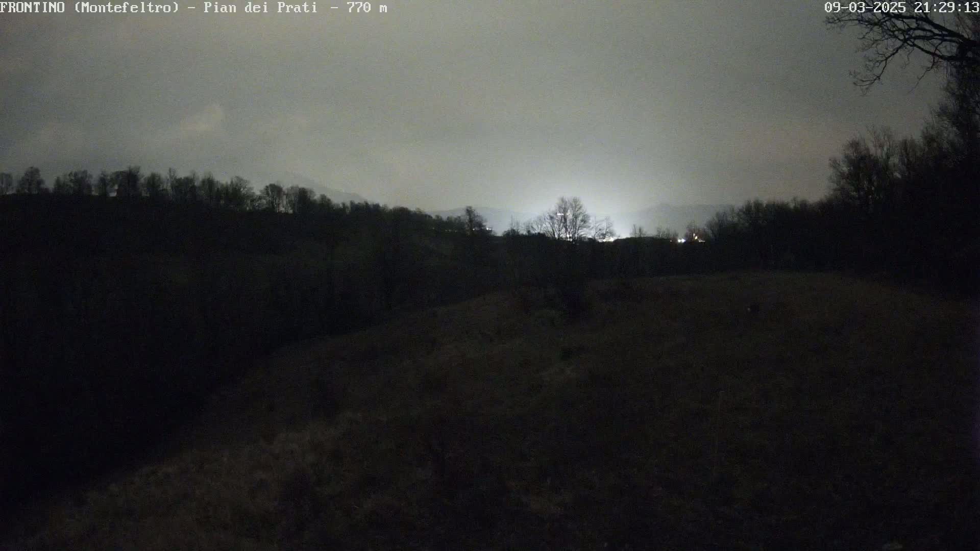 A nighttime view of a dark, hilly landscape under a hazy, overcast sky, with a distant area illuminated by artificial light.