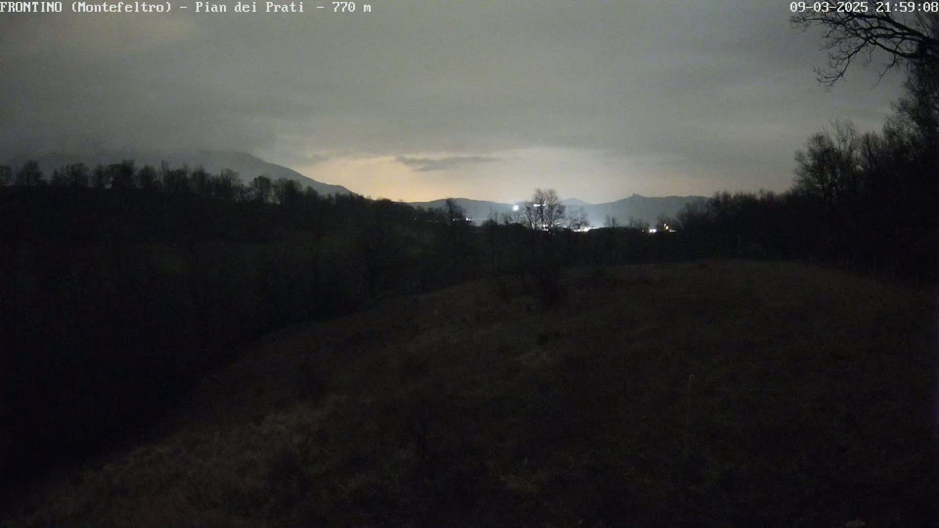 A nighttime view of a dark, hilly landscape under a cloudy sky, with distant lights faintly visible in the background.