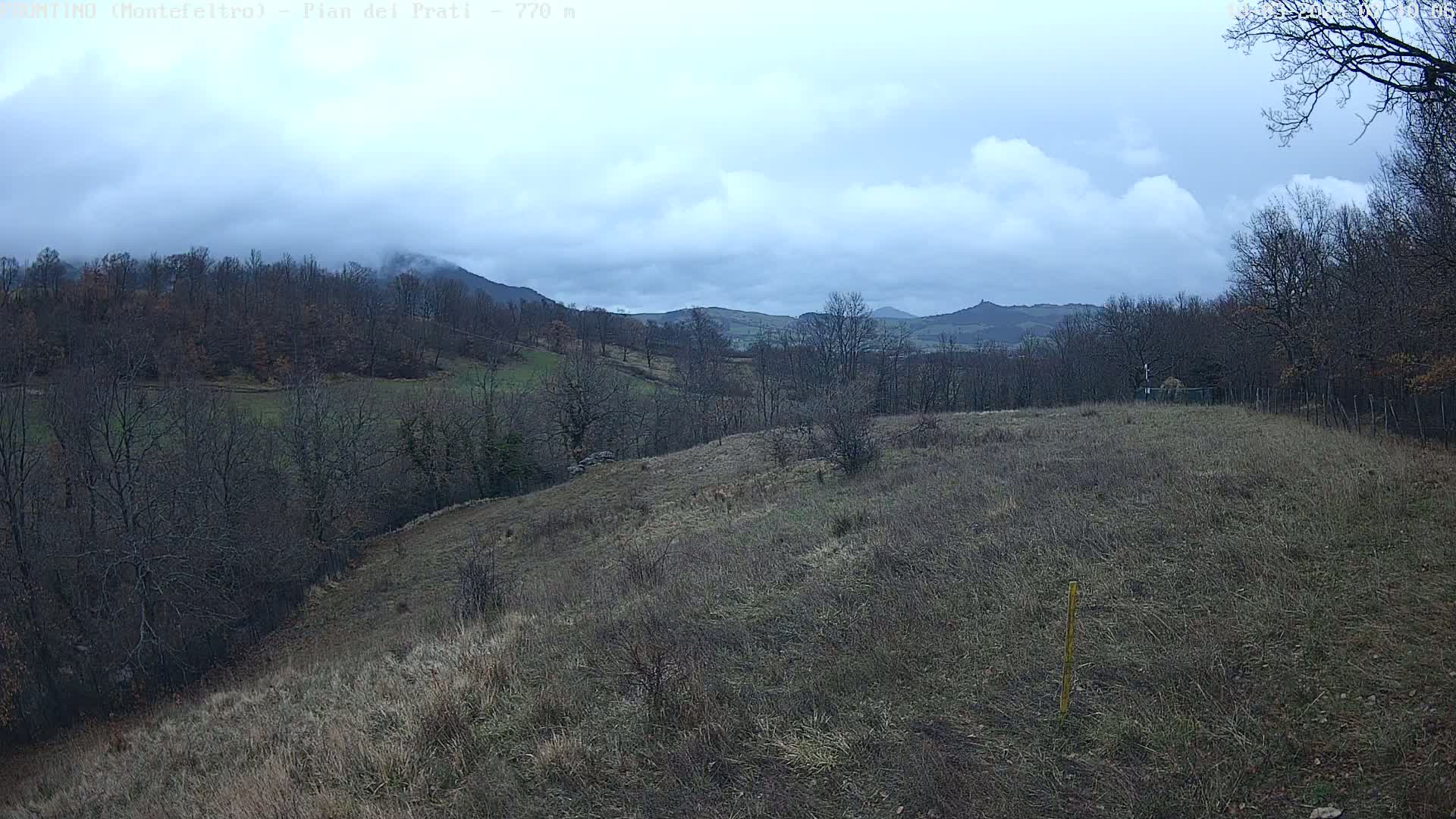 A mostly cloudy day reveals a hilly landscape with leafless trees and brown, grassy fields extending to distant mountains.
