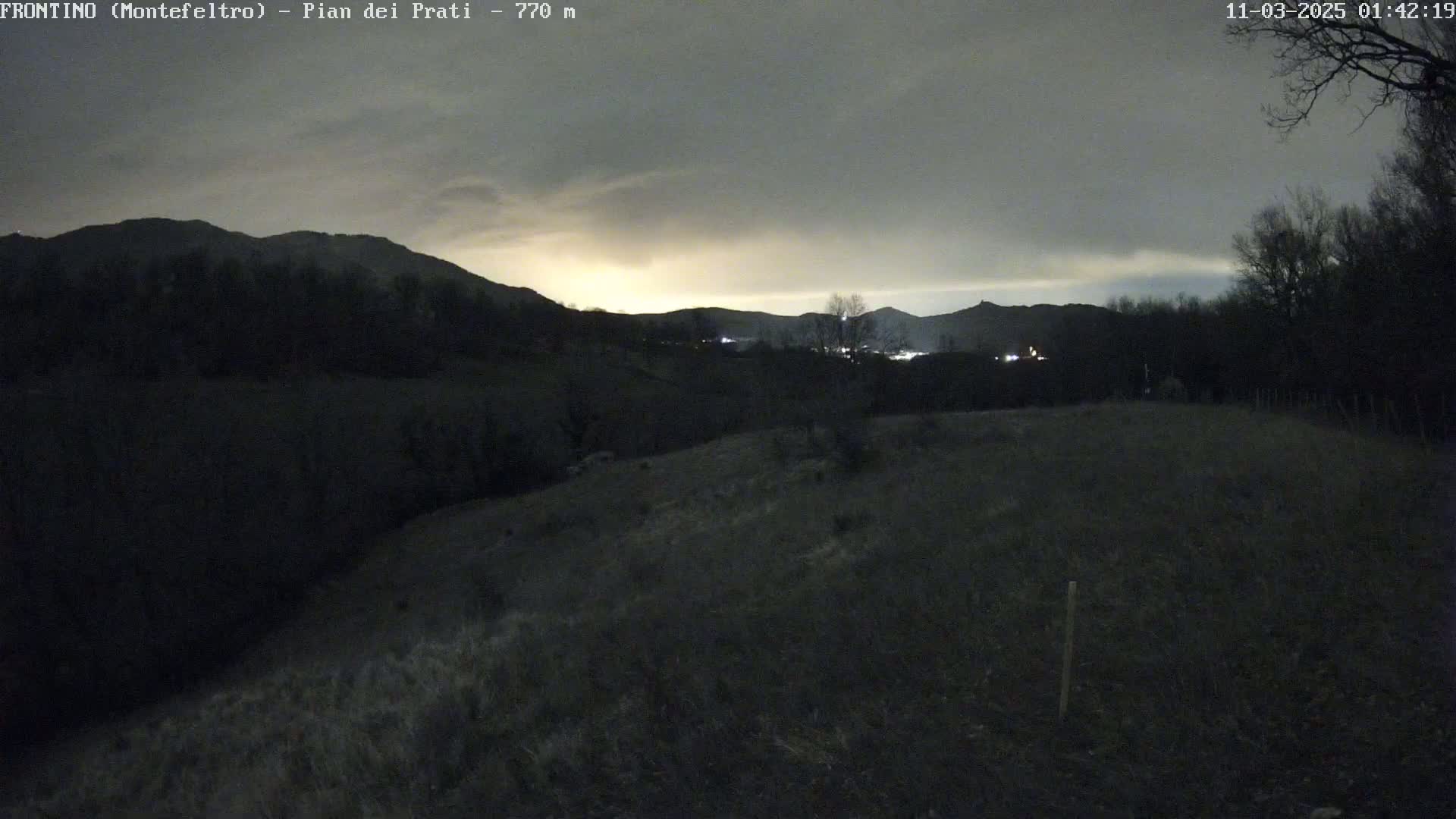 A dark, overcast night scene shows a hilly landscape with sparse vegetation under a cloudy sky, faintly illuminated by distant lights.