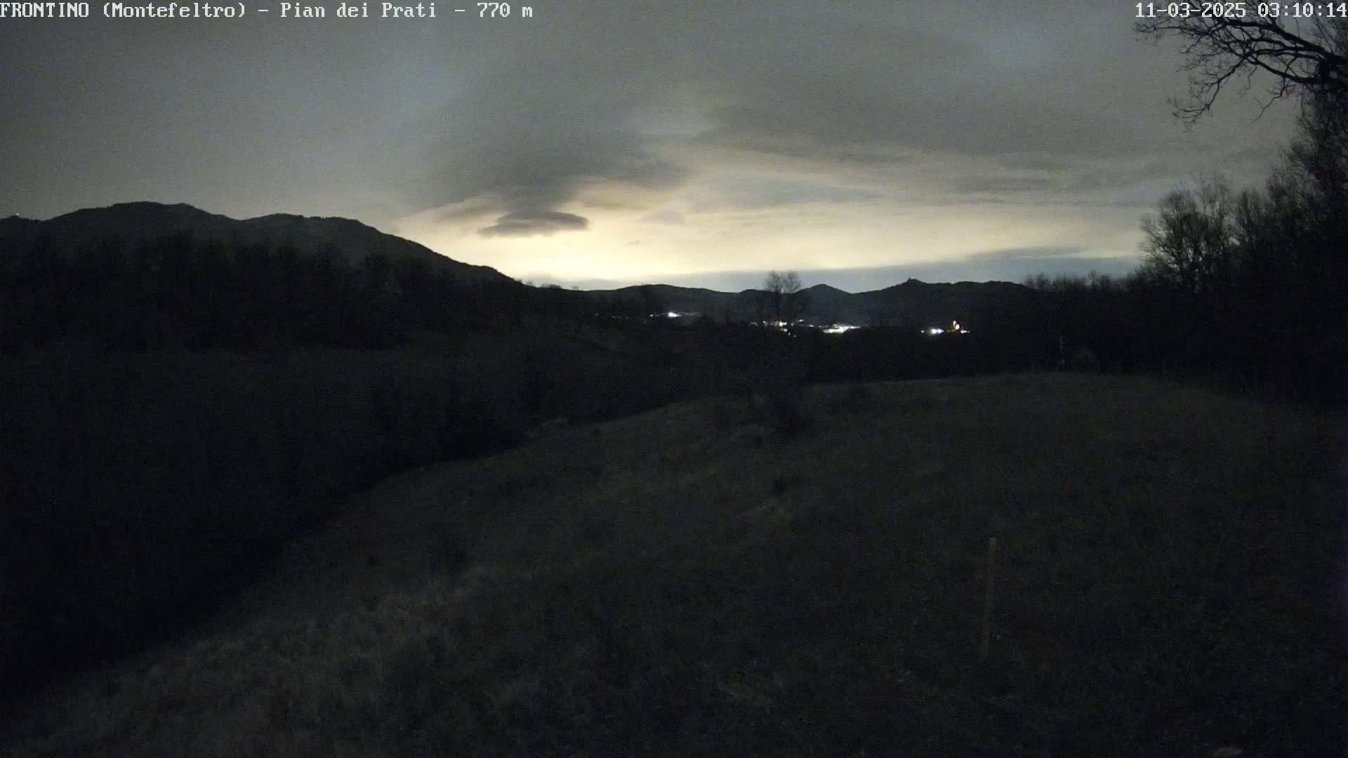 A dark, overcast night scene shows rolling hills and distant lights under a pale, cloudy sky.