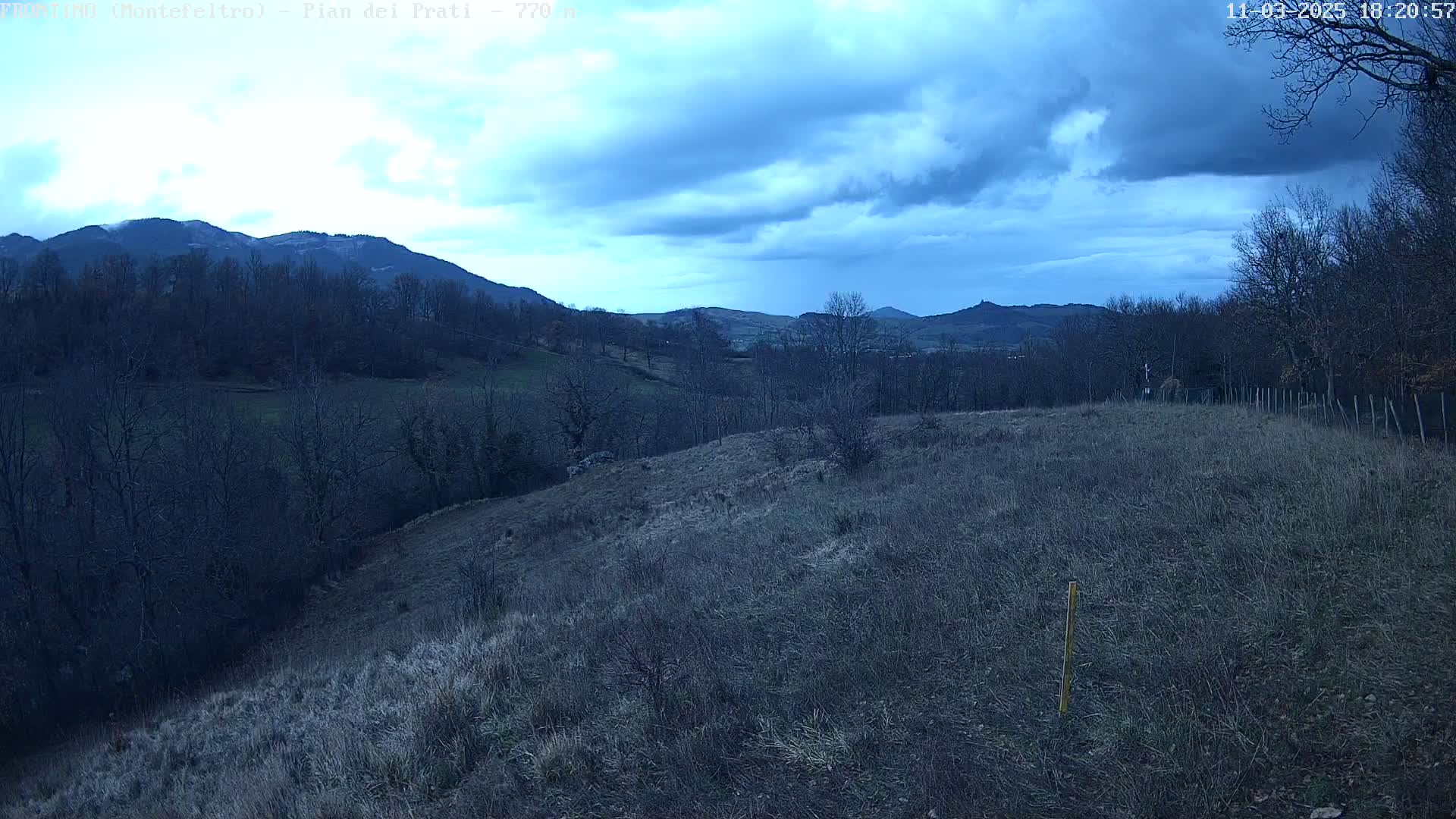 A view of a hilly landscape under a cloudy, twilight sky shows leafless trees and brown, grassy fields.