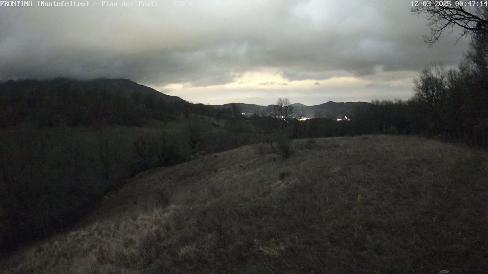 A dark, overcast sky hangs over a hilly landscape of dark green trees and brown, grassy fields.