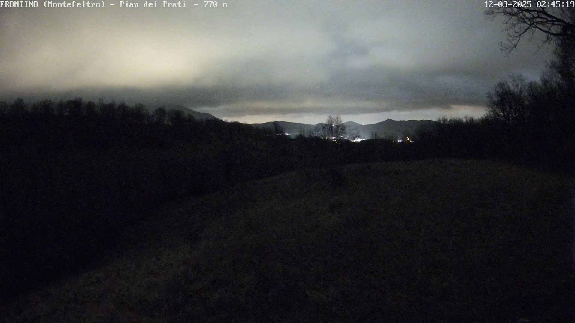 A nighttime, overcast view of dark hills and distant, faintly lit towns under a cloudy sky.