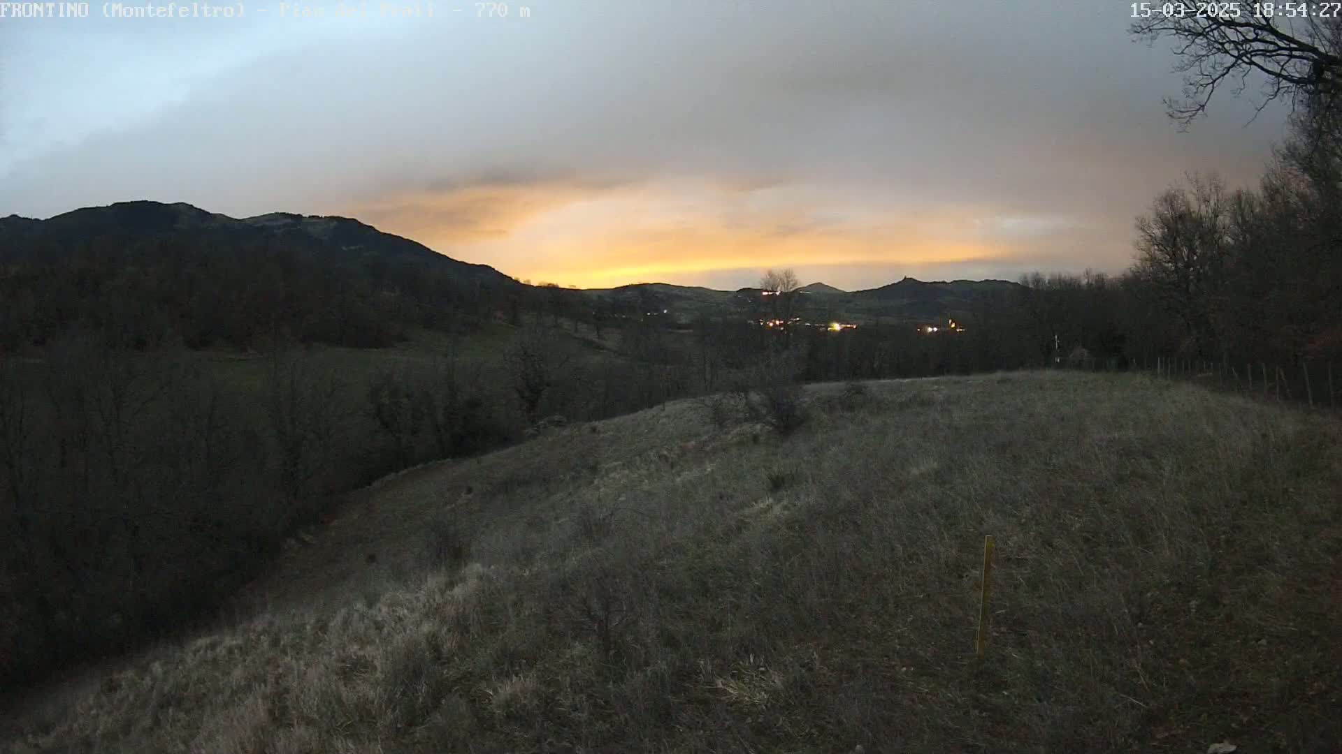 A hilltop view at dusk shows a valley with a town and distant mountains under a cloudy sky with a sunset.