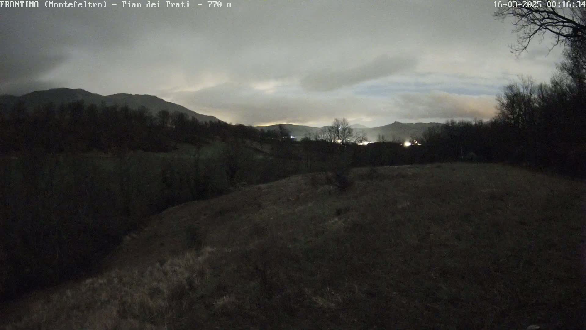 A dark, overcast night scene shows a hilly landscape with sparse, leafless trees and distant lights under a cloudy sky.