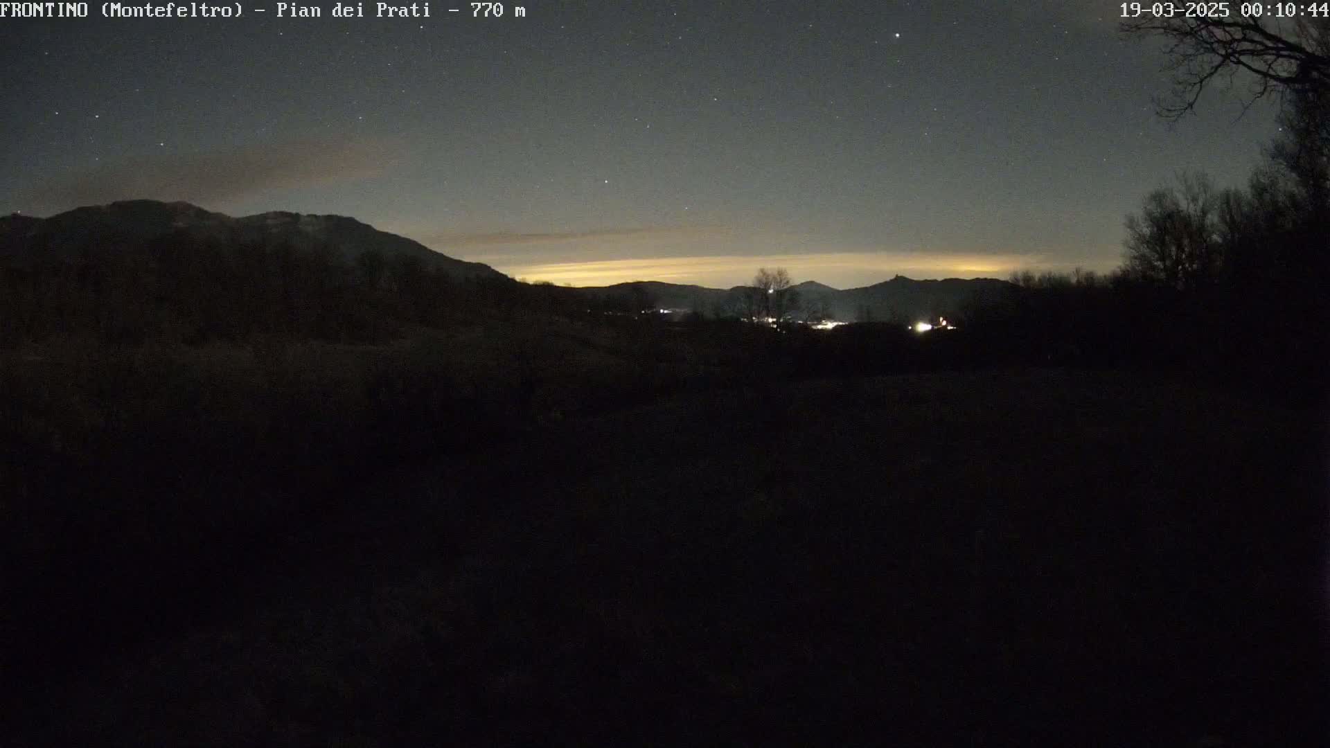 A nighttime view of dark, silhouetted mountains and a valley with a few lights visible under a mostly clear, starry sky.