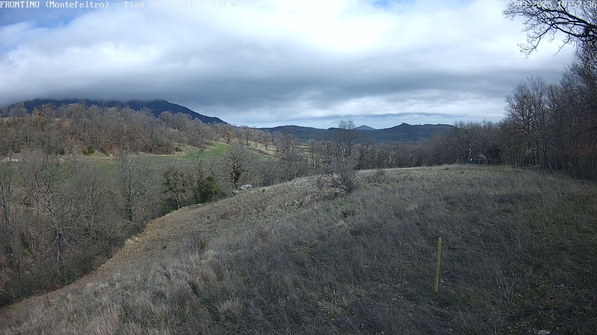 A mostly cloudy day reveals a hilly landscape of leafless trees and dry, brown fields, with mountains in the distance.