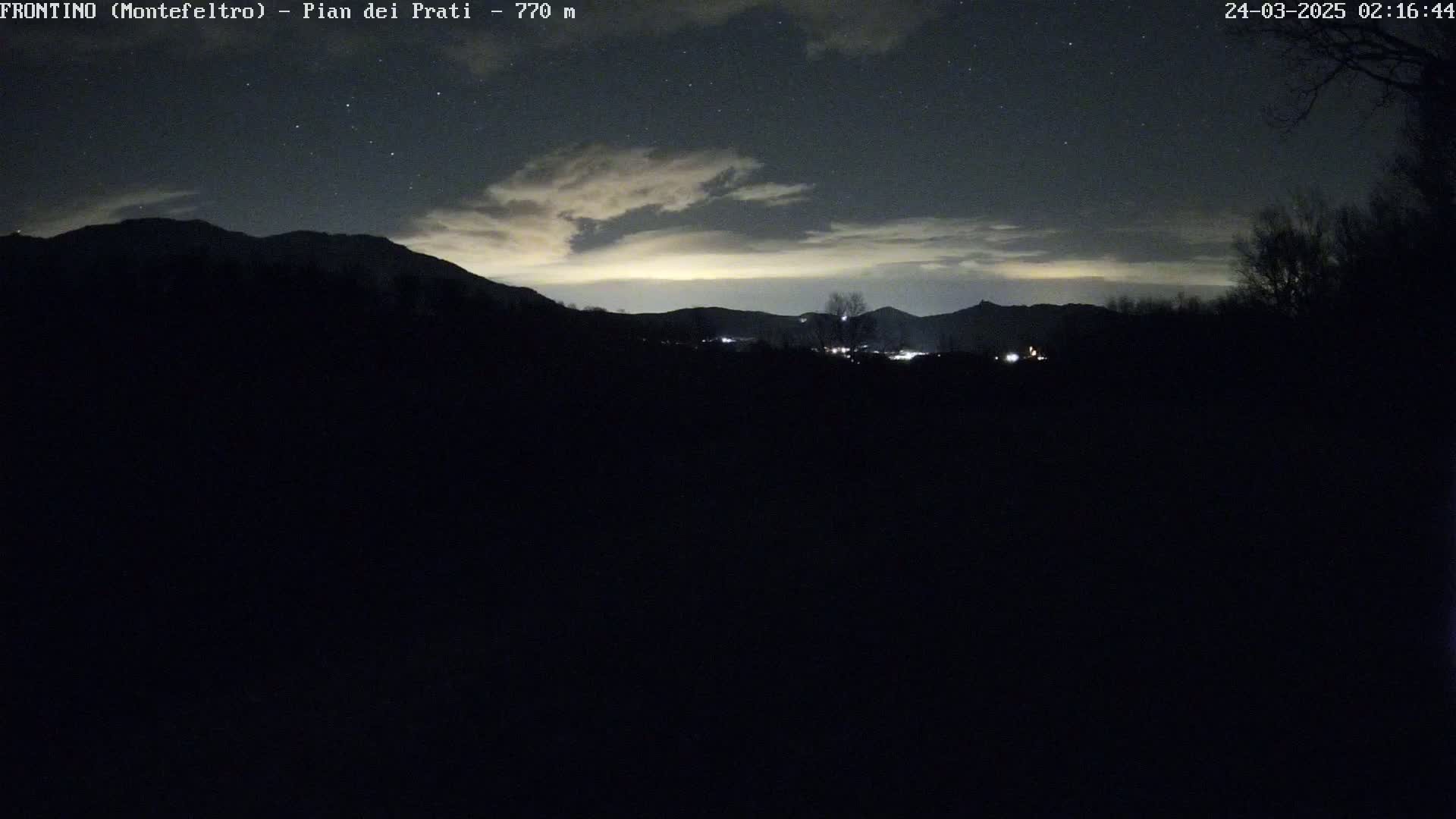 A nighttime view of a distant, dimly lit town nestled in a valley between silhouetted mountains under a partly cloudy sky dotted with stars.