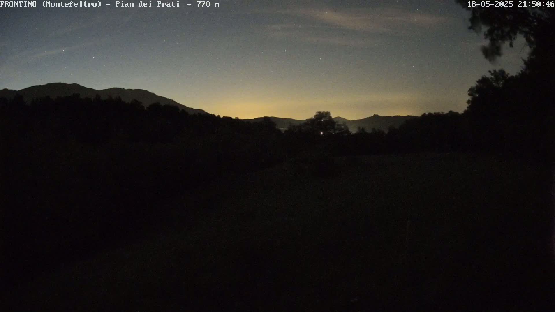 A nighttime, starlit view of dark, silhouetted mountains and trees under a mostly clear sky with a faint, pale yellow glow on the horizon.