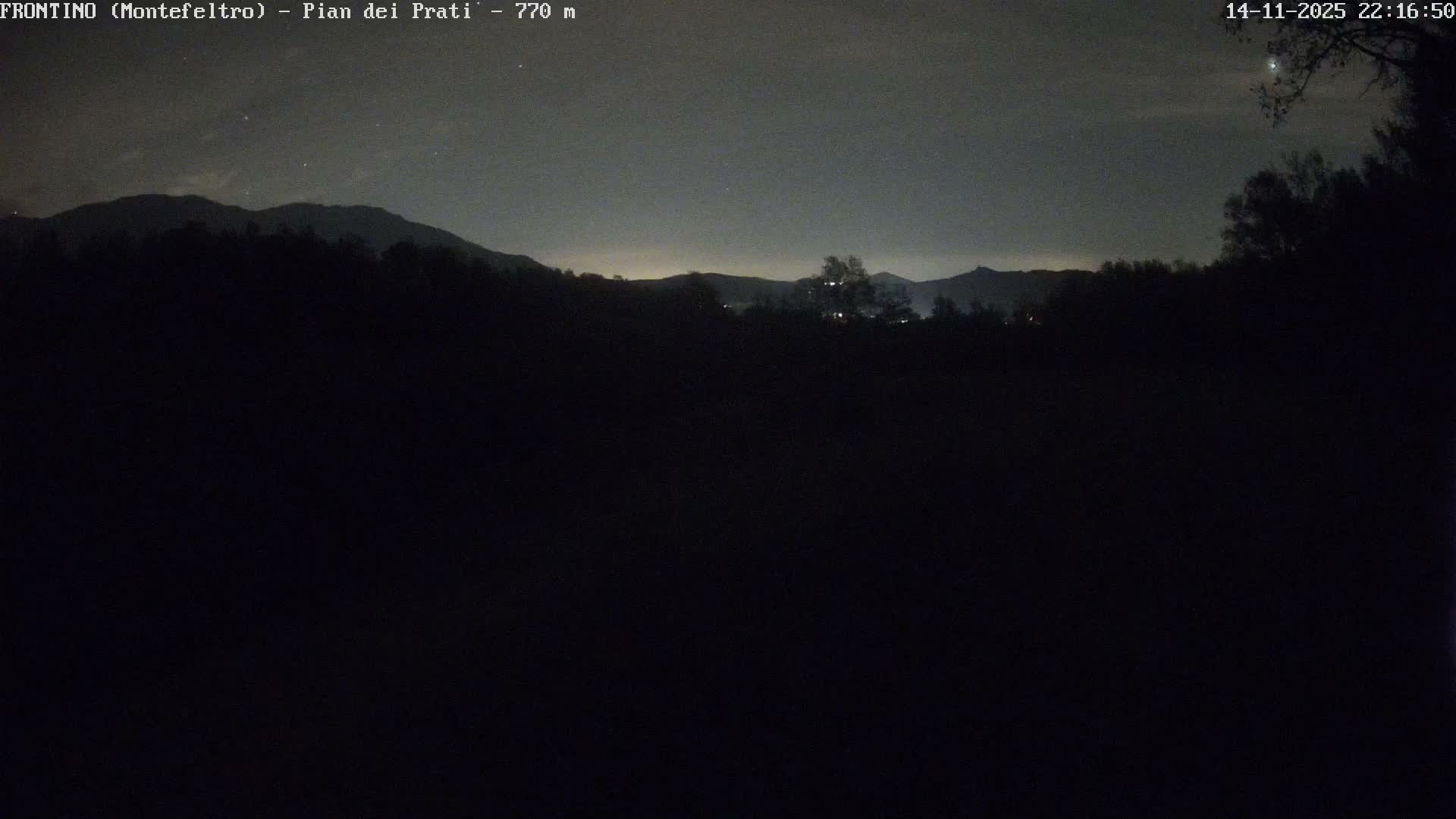A dark nighttime landscape reveals silhouetted trees and mountains under a partly cloudy sky, with a faint glow of distant lights on the horizon and two bright celestial objects visible in the upper right.