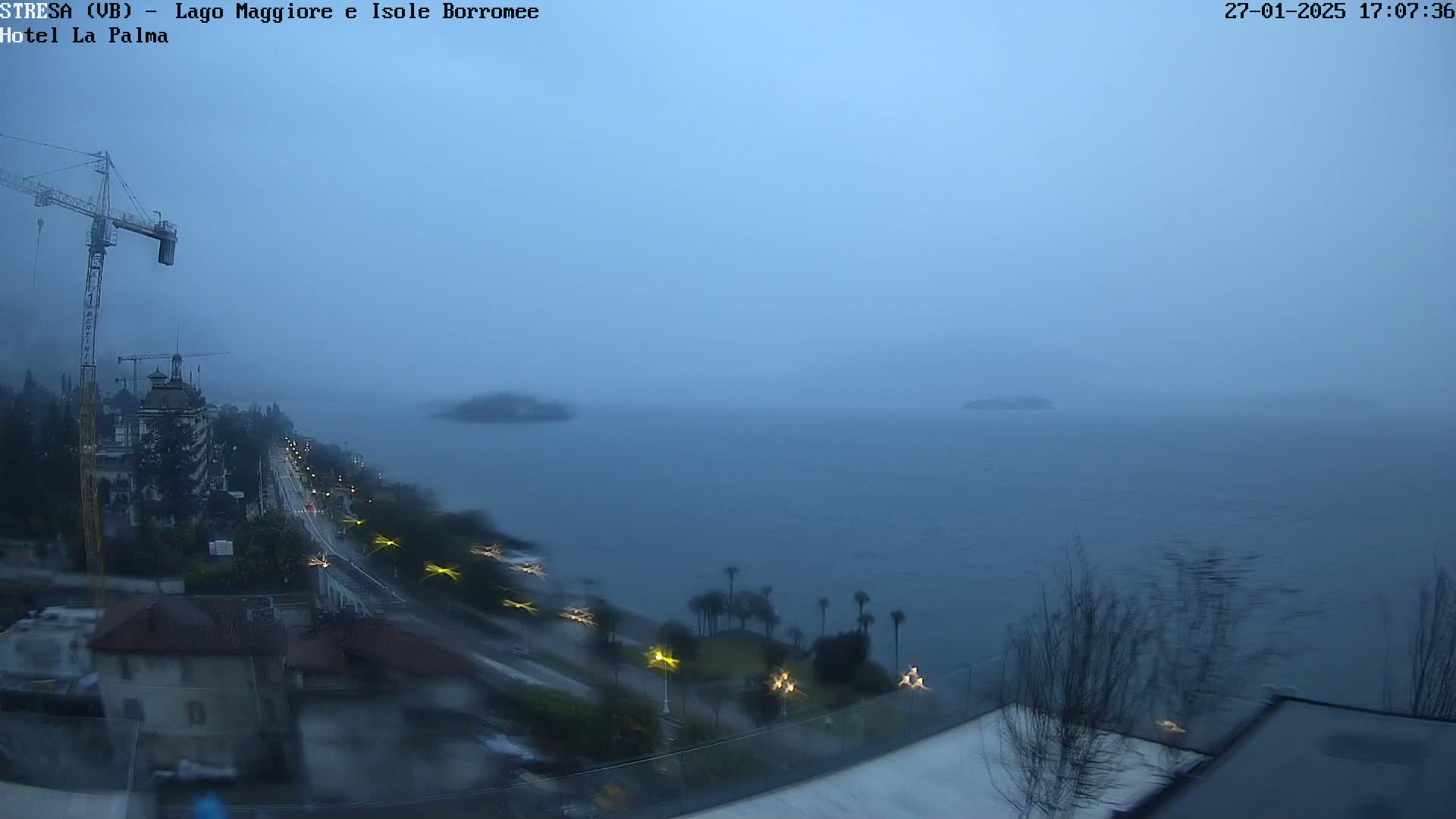 A hazy, rainy view of a large body of water with islands in the distance, a shoreline with buildings and a road, and a construction crane visible.