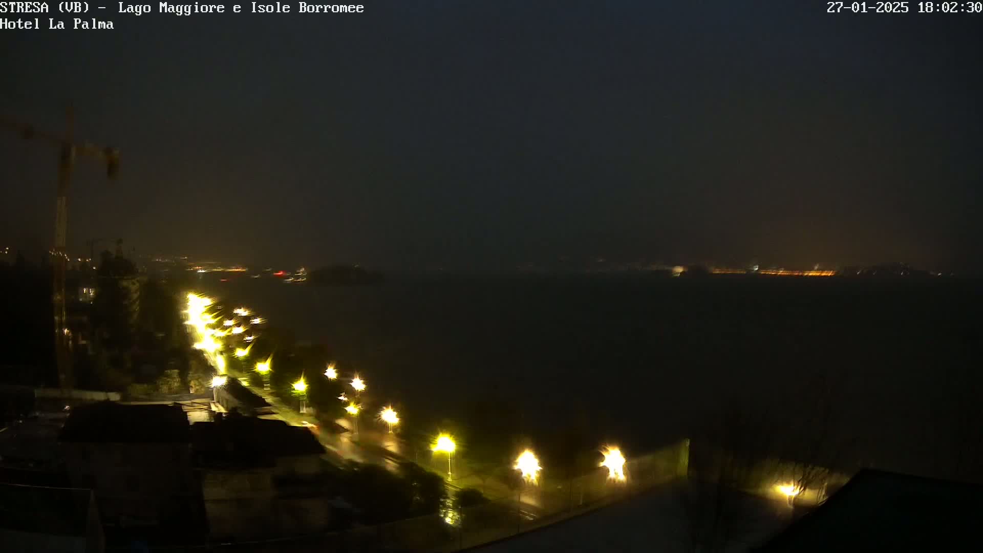 A nighttime view of a rain-soaked city overlooking a dark body of water, illuminated by streetlights along a road curving toward the water.