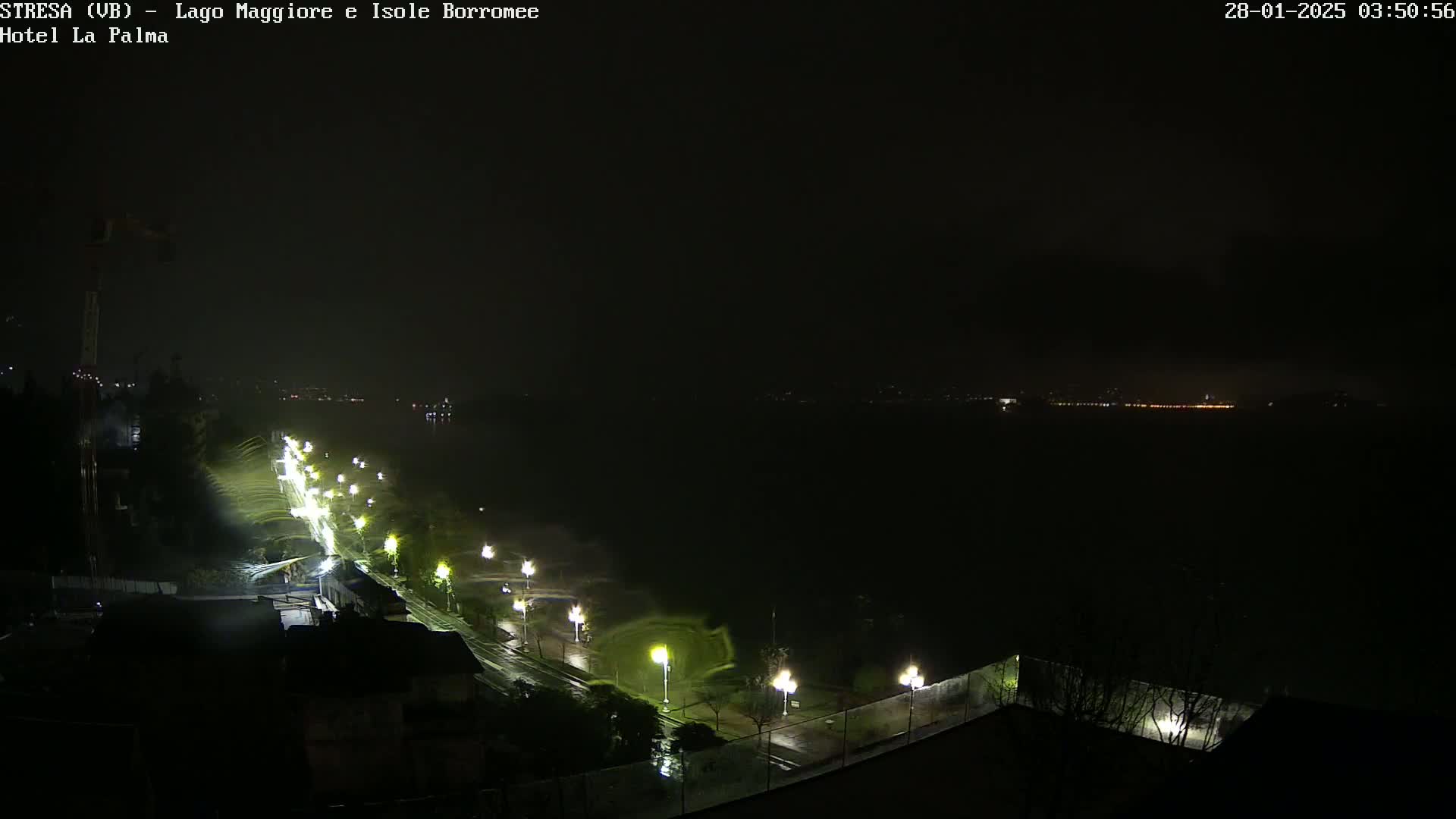 A nighttime, rain-swept view of a shoreline road illuminated by streetlights, with a dark body of water and distant city lights visible in the background.
