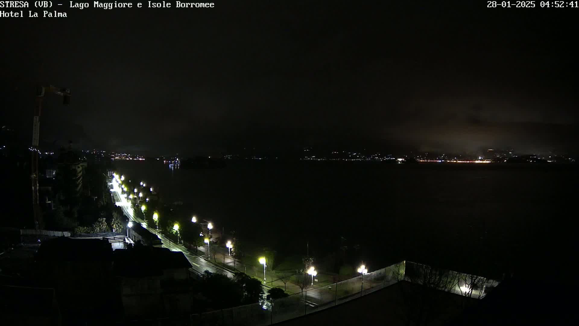 A nighttime, overcast view of a dark body of water with a city skyline faintly visible in the distance, and a lit road curving along the waterfront in the foreground.