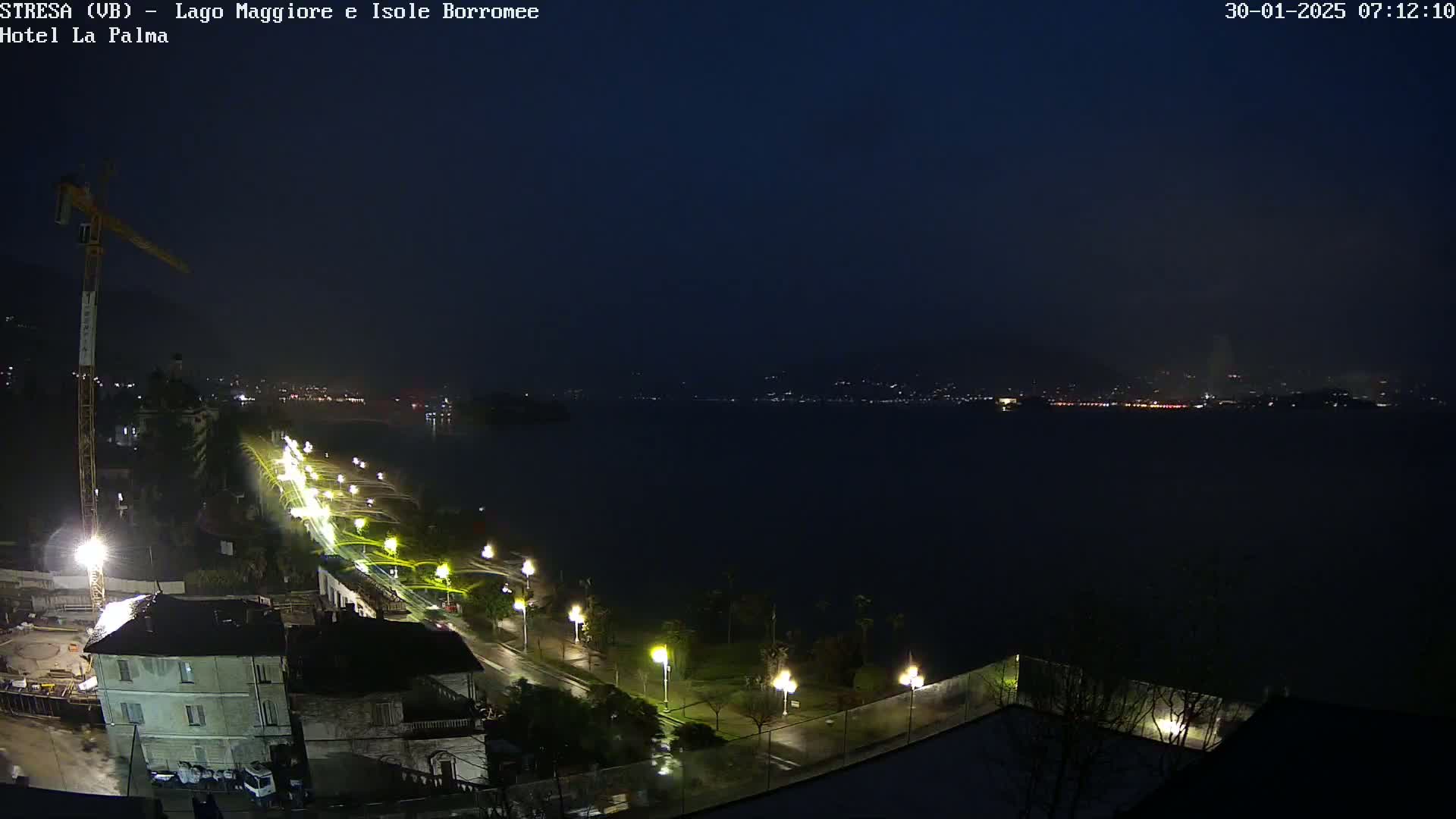 A nighttime view of a lakeside town with a construction crane, streetlights illuminating a wet roadway, and a dark body of water with a distant, faintly lit cityscape under a dark, possibly overcast sky.
