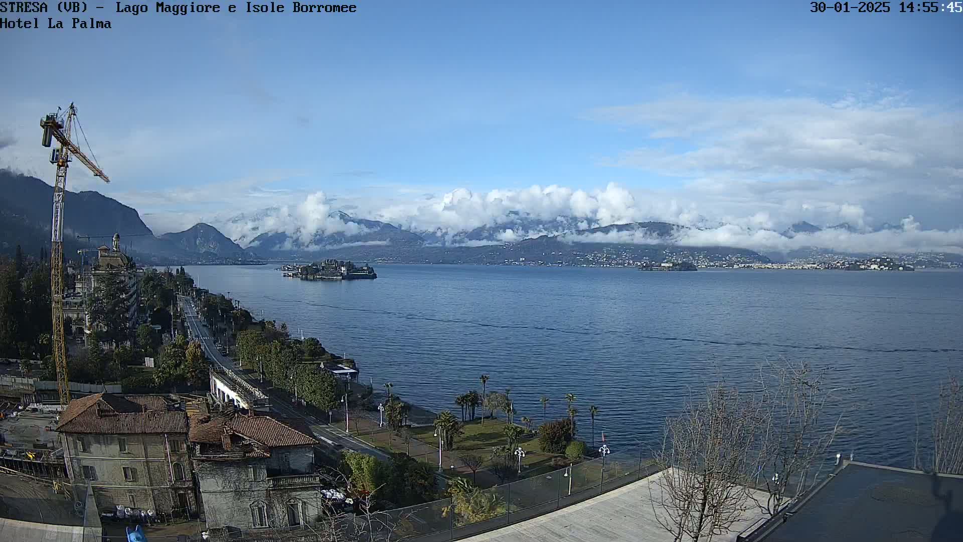 A partly cloudy day overlooks a large body of water with mountains in the background, a shoreline with buildings and vegetation, and a construction crane visible nearby.