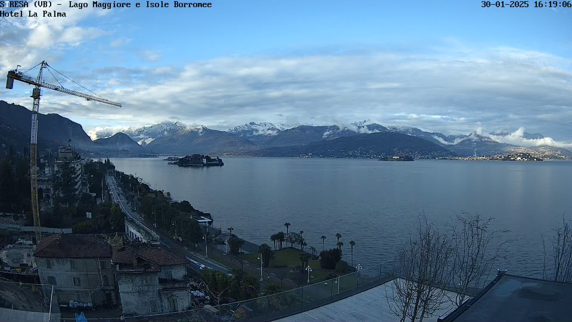 A partly cloudy day reveals a large lake with snow-capped mountains in the distance, a small island in the middle, and a shoreline with buildings and a construction crane.