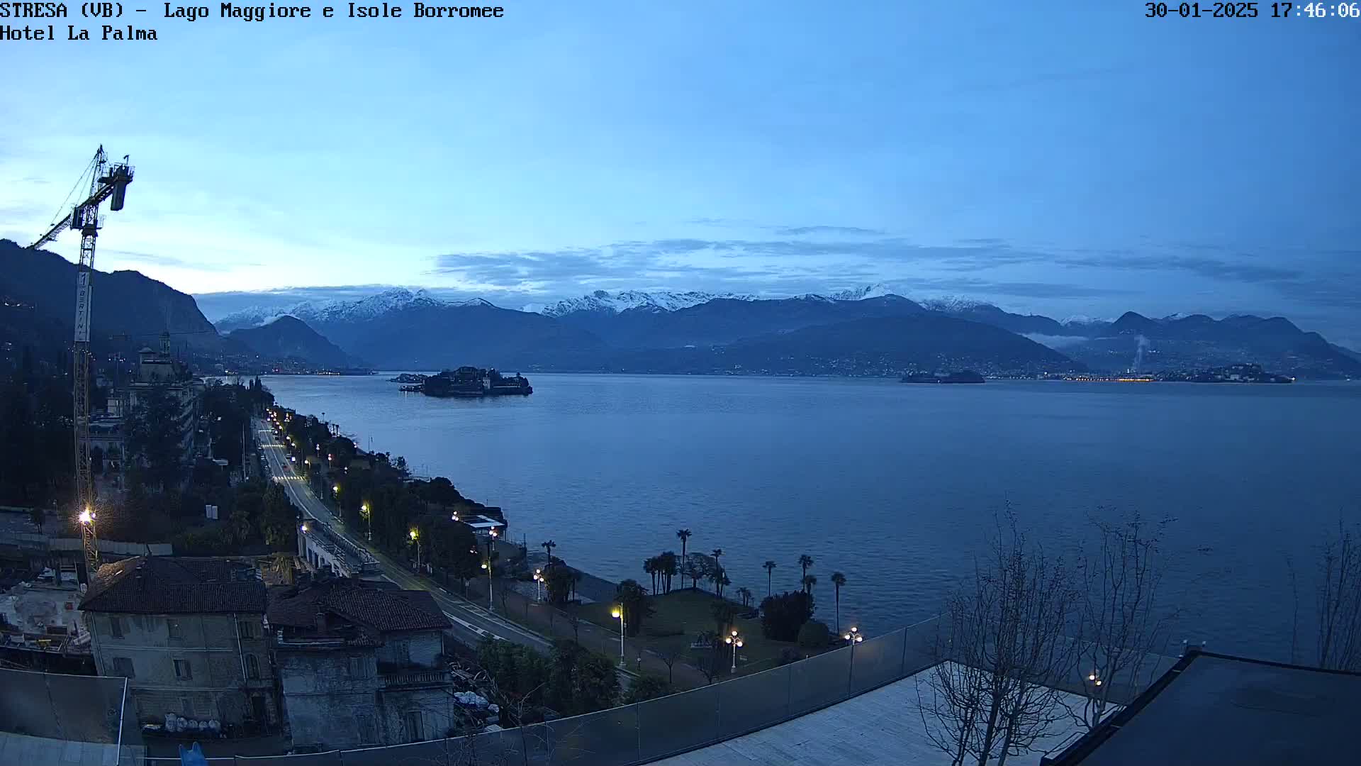 A twilight view of a lake with snow-capped mountains in the background, a shoreline with buildings and a road, and a construction crane visible near the foreground.