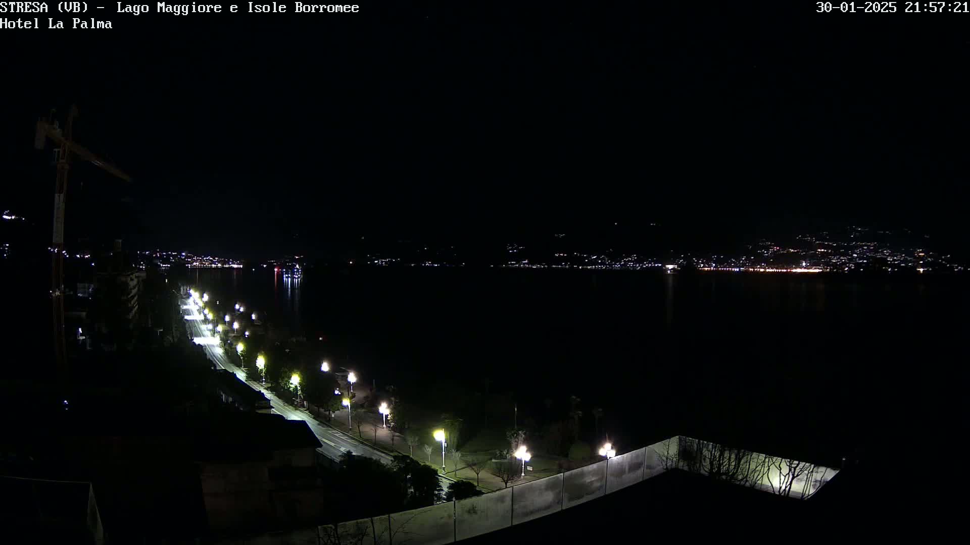 A nighttime view of a city across a dark body of water, with a lit road curving along the shoreline toward the viewer.