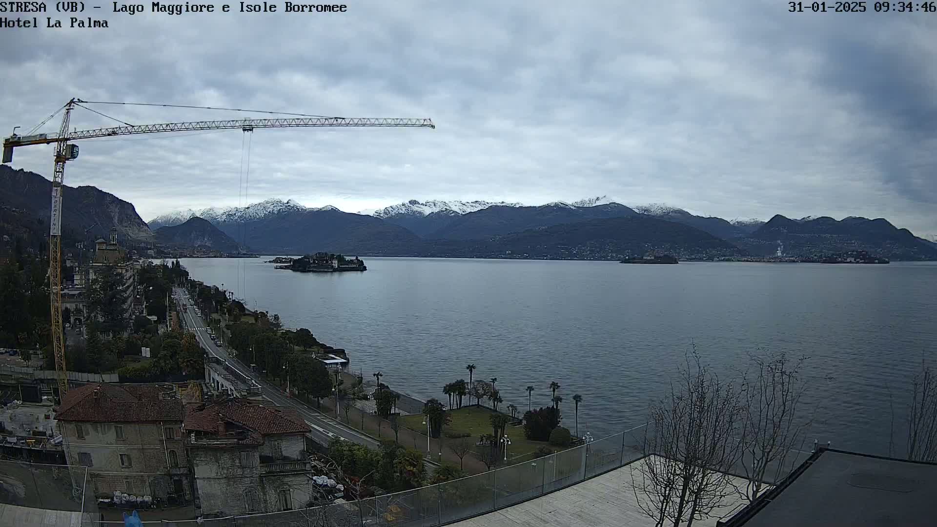 A large body of water is seen with snow-capped mountains in the background, a small island in the middle, and a construction crane on the left, all under an overcast sky.