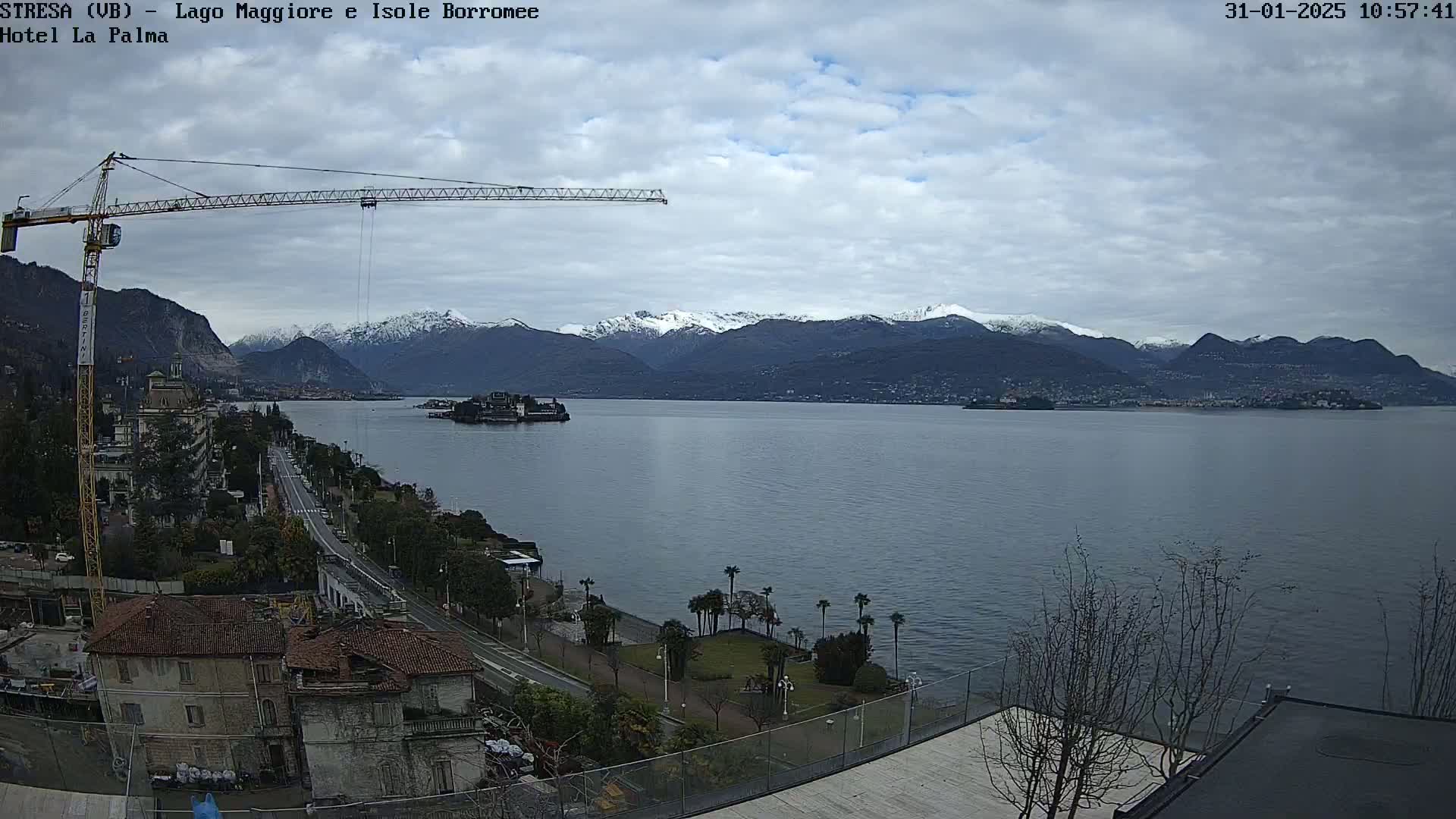 A large body of water stretches towards snow-capped mountains under a cloudy sky, with a crane visible near a shoreline featuring buildings and palm trees.