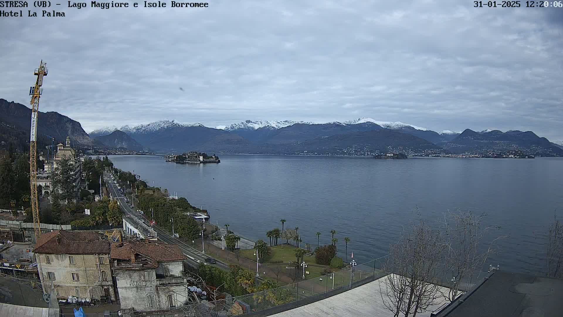 A lakefront view shows a town with buildings, a road, and a crane, overlooking a calm lake with a small island and snow-capped mountains in the distance under a cloudy sky.