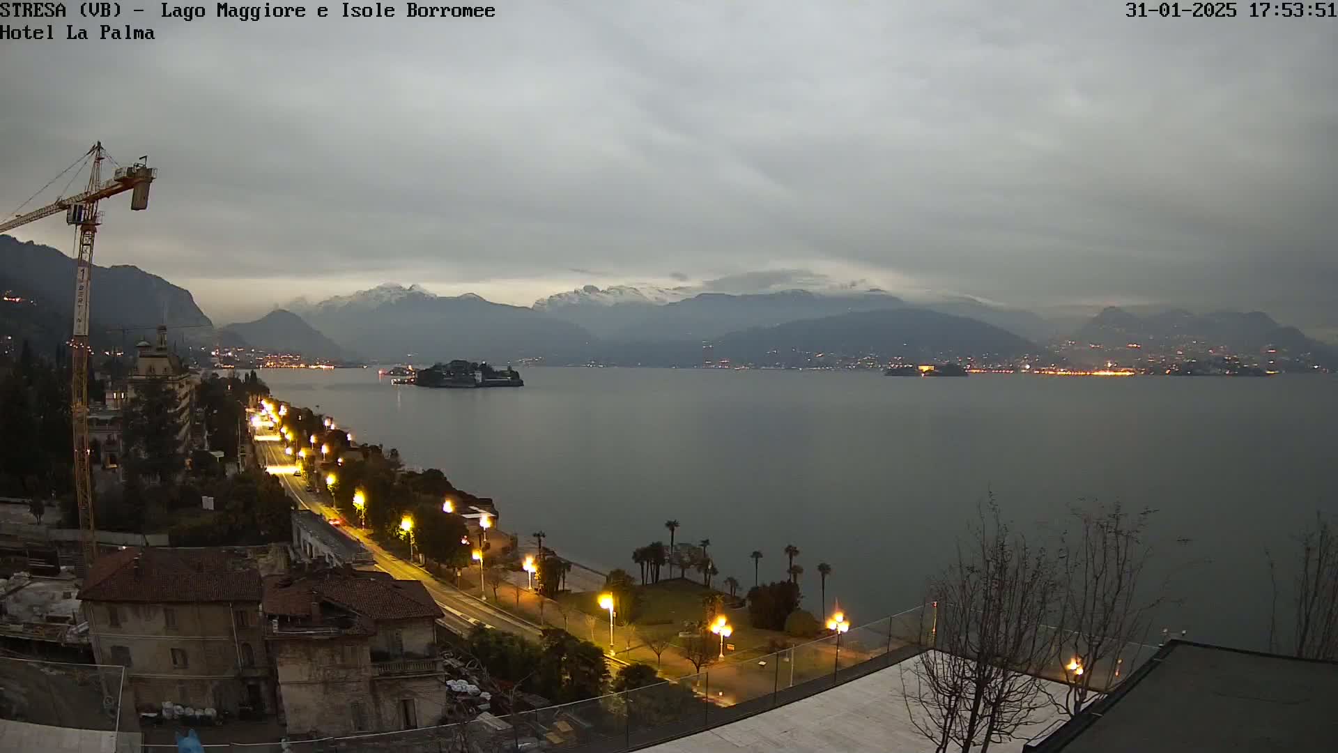 A nighttime view of a calm lake with snow-capped mountains in the distance, under an overcast sky, features a lakeside road lined with lights and buildings along the shore.
