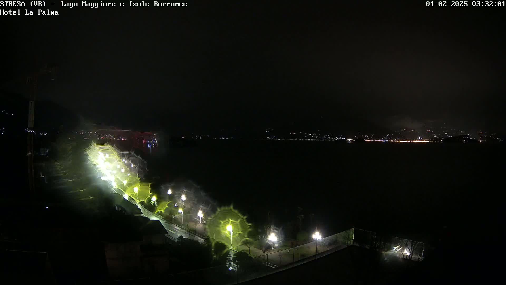 A nighttime, blurry view of a lakeside town with streetlights illuminating a path along the water's edge under a dark, possibly stormy sky.