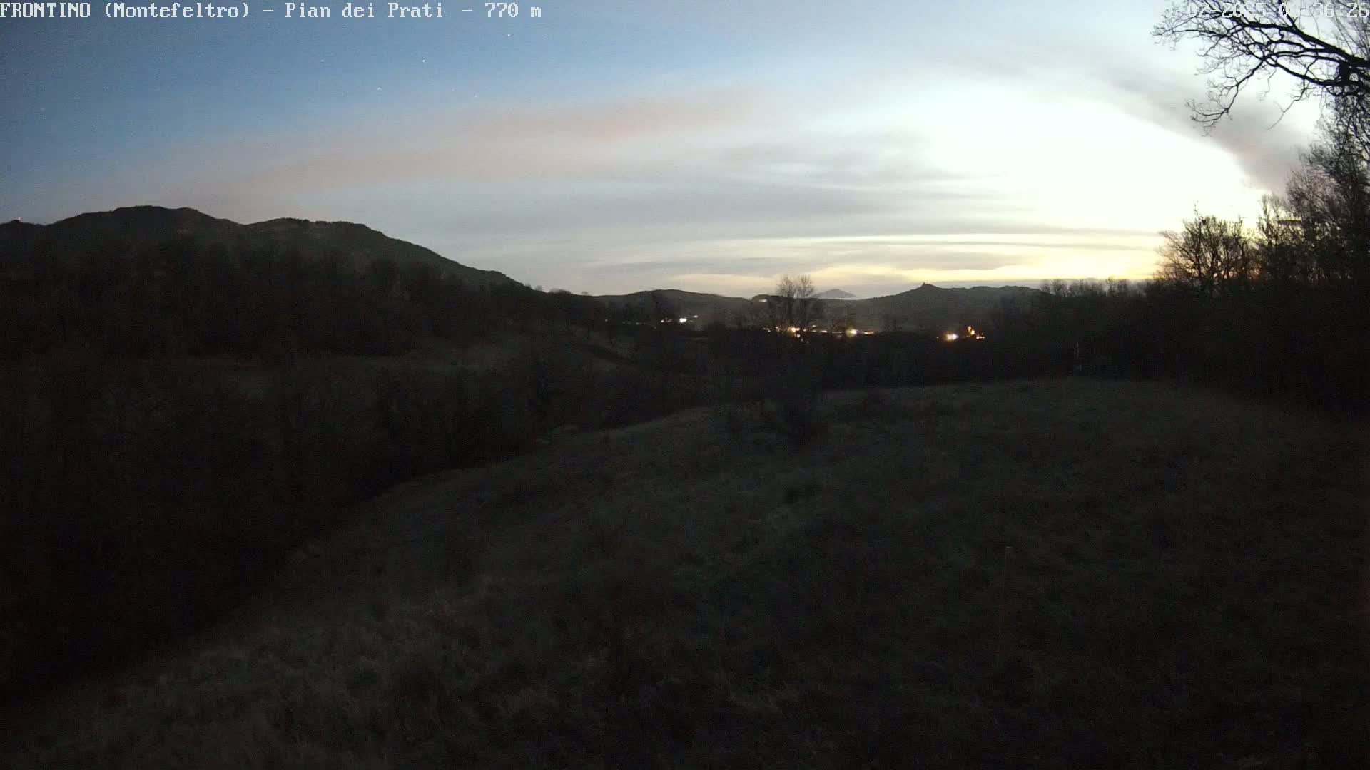 A dark, grassy field stretches out before a line of silhouetted trees and hills under a twilight sky.
