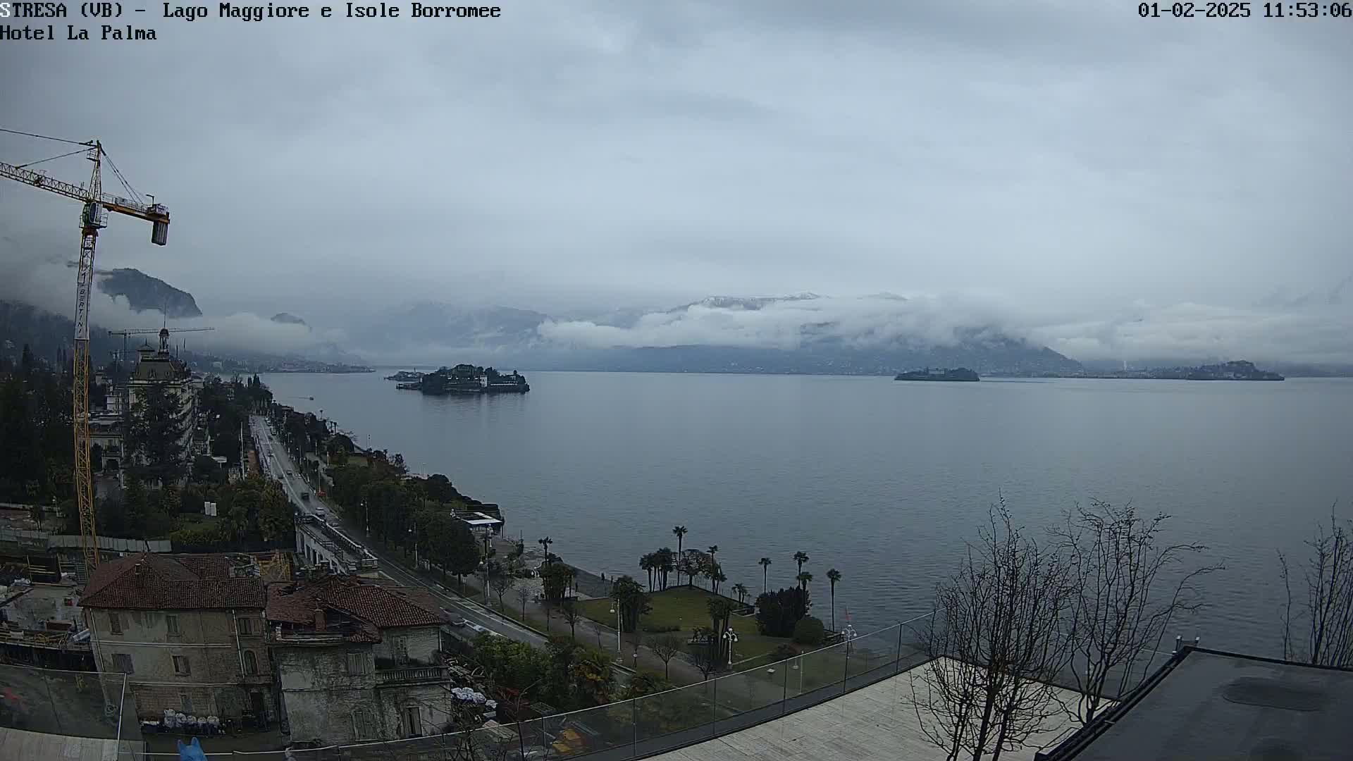 A large body of water stretches to snow-capped mountains under a cloudy sky, with a lakeside town, palm trees, and a construction crane visible in the foreground.