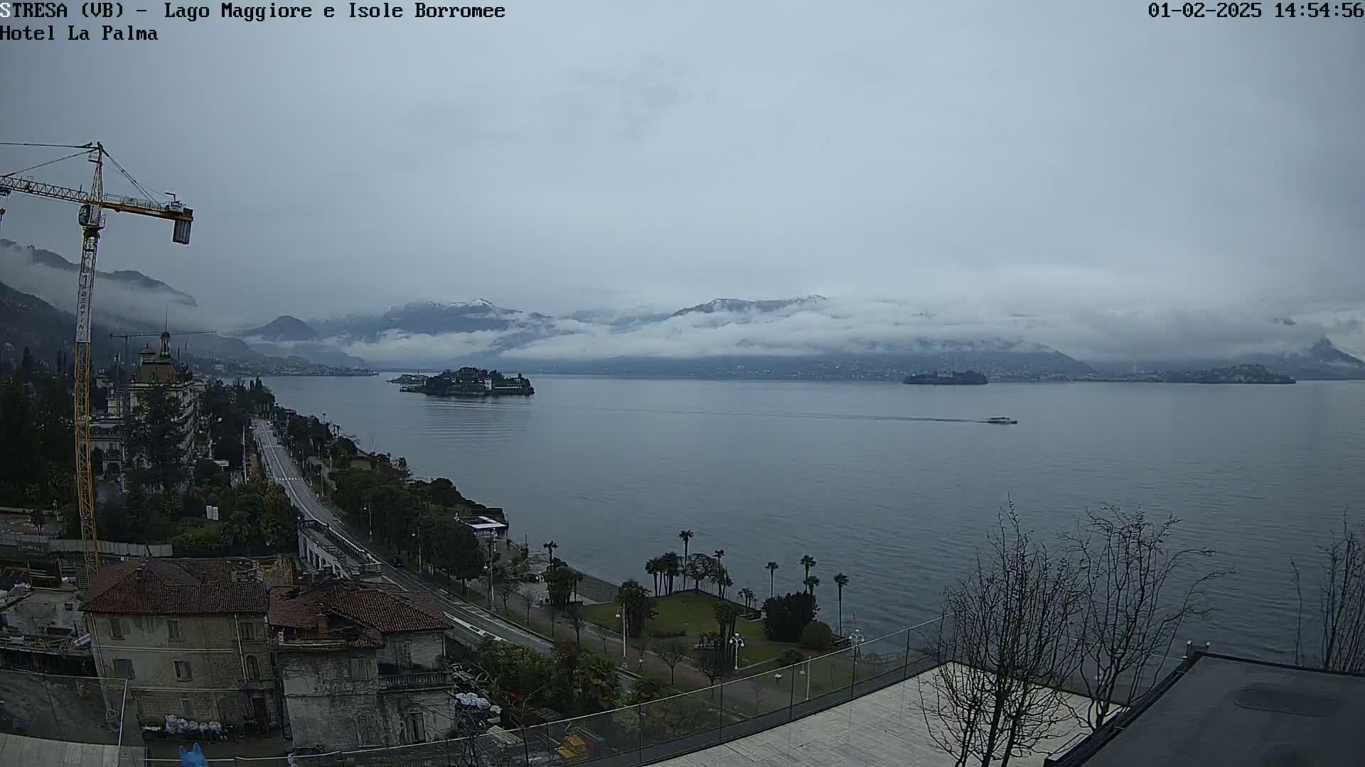 A cloudy day overlooking a large lake with snow-capped mountains in the distance, a small island, and a construction crane visible in the foreground.