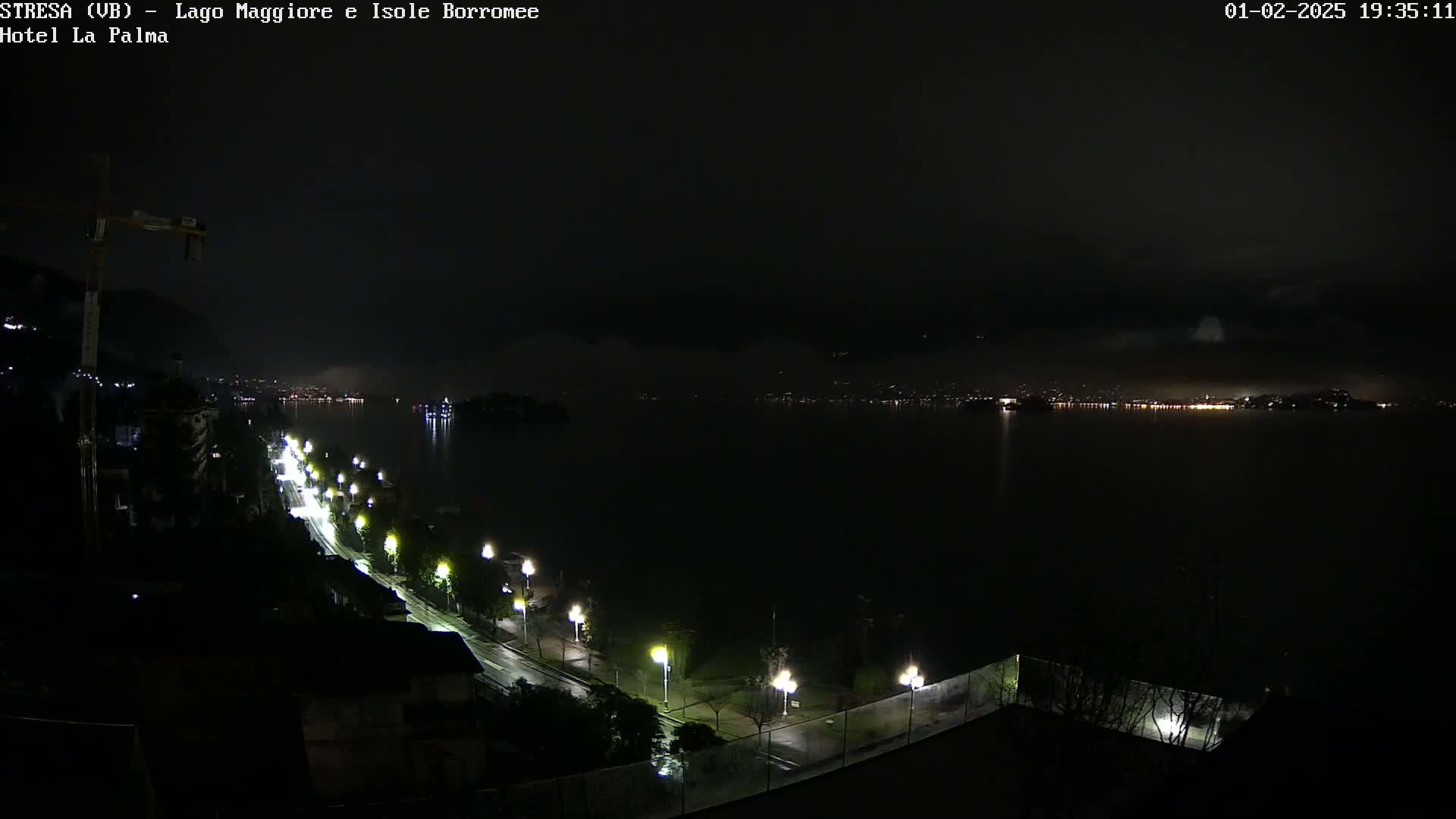 A rain-slicked road, lined with lit streetlights, curves alongside a dark body of water reflecting the lights of a distant city at night.