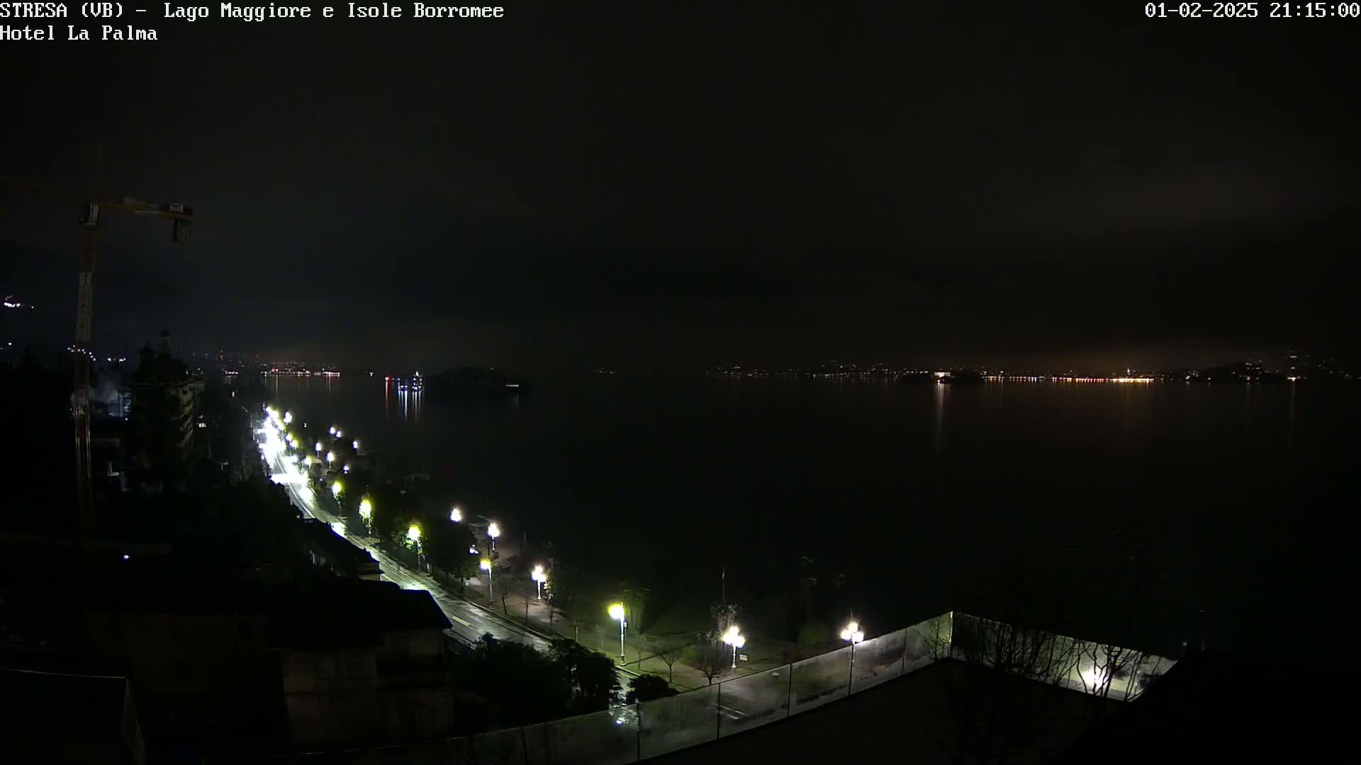 A nighttime view of a dark, calm lake with a shoreline road lit by streetlights under a cloudy sky.