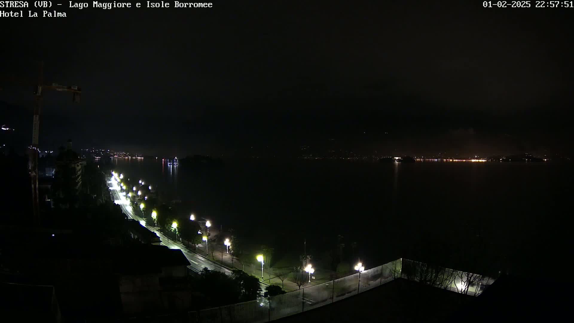 A nighttime, rain-slicked road lined with lights curves along a dark lake shore, with a distant city glowing faintly across the water.